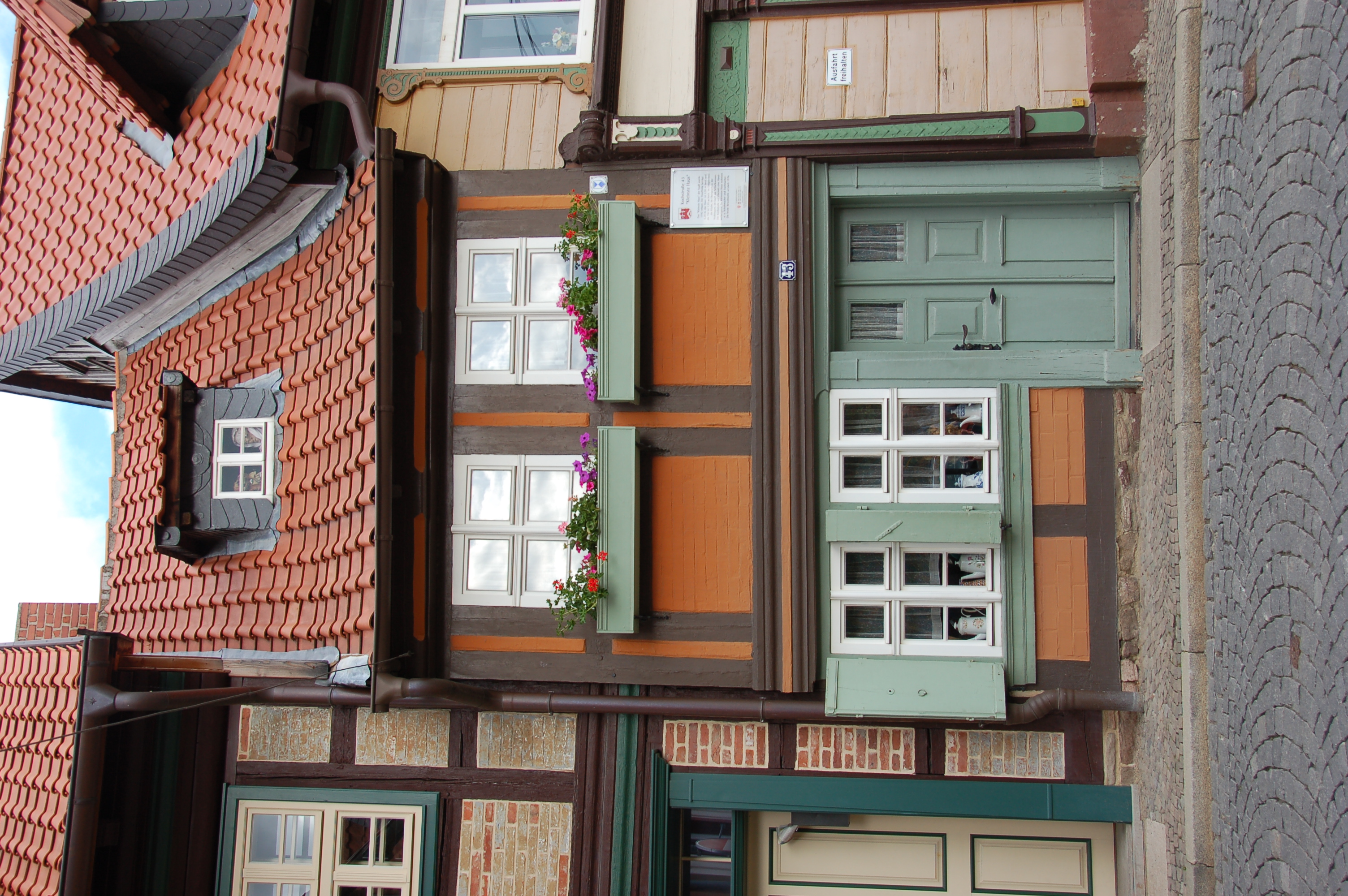 photograph shows a small half-timbered house, sitting between two taller houses. The framing is painted brown and the infill orange. Door, shutters and window boxes in front of the first floor windows are painted in a pastel green. There are flowers in the window boxes and white china coffee pots are displayed in the ground floor windows.