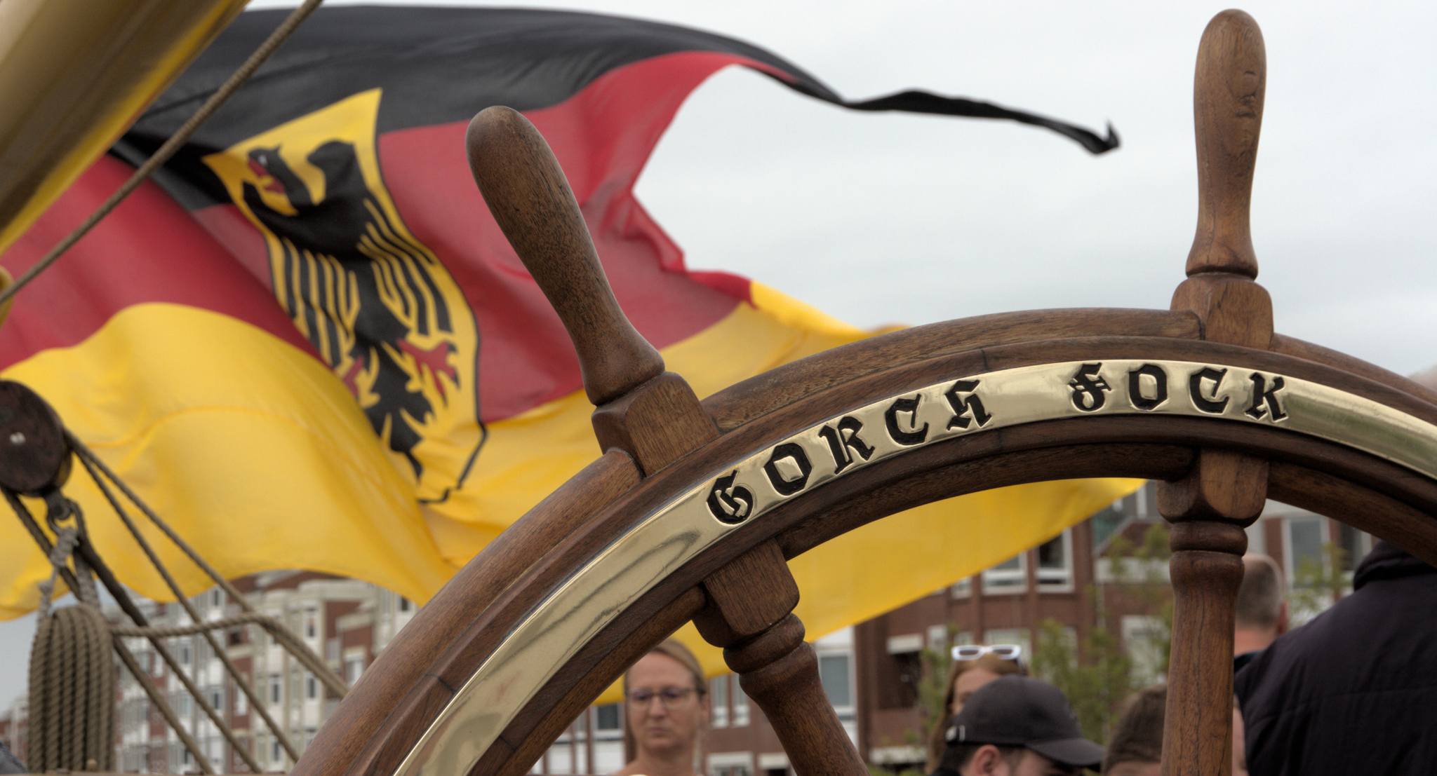 wooden steering wheel of the sailing ship Gorch Fock, with an inset brass ring reading "Gorch Fock", and the flying German jack in the background