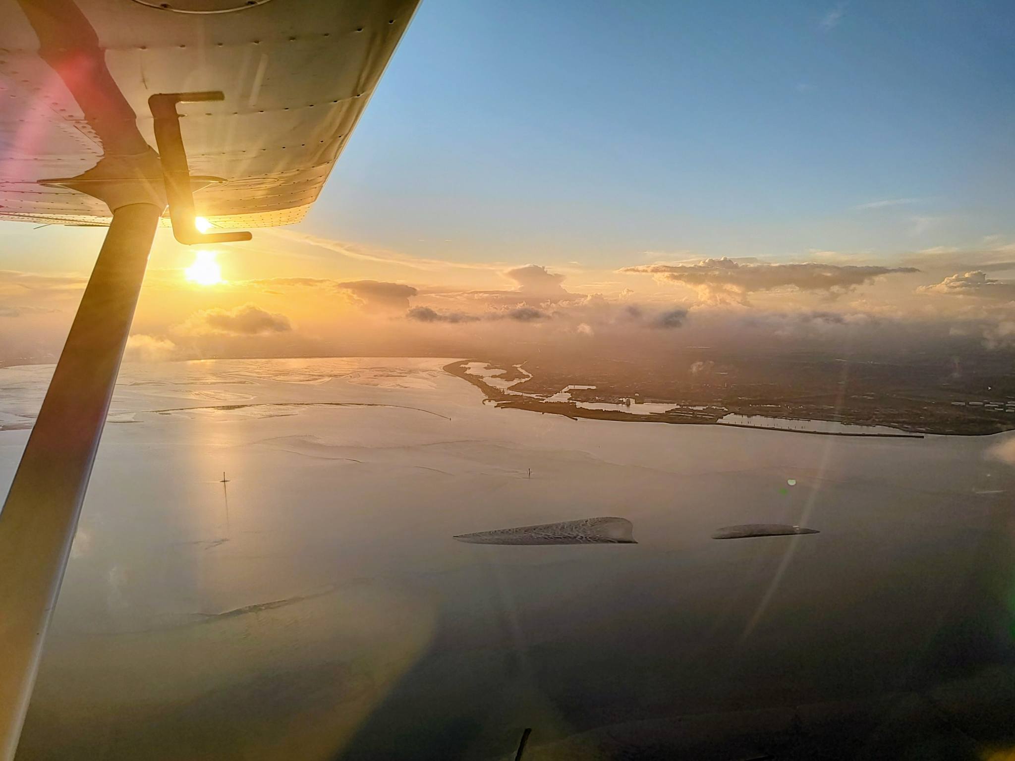 aerial view of the Jade Bight and Wilhelmshaven along the left wing of a Tecnam P2010, flying at 2500 feet; in the background the setting sun is illuminating some clouds
