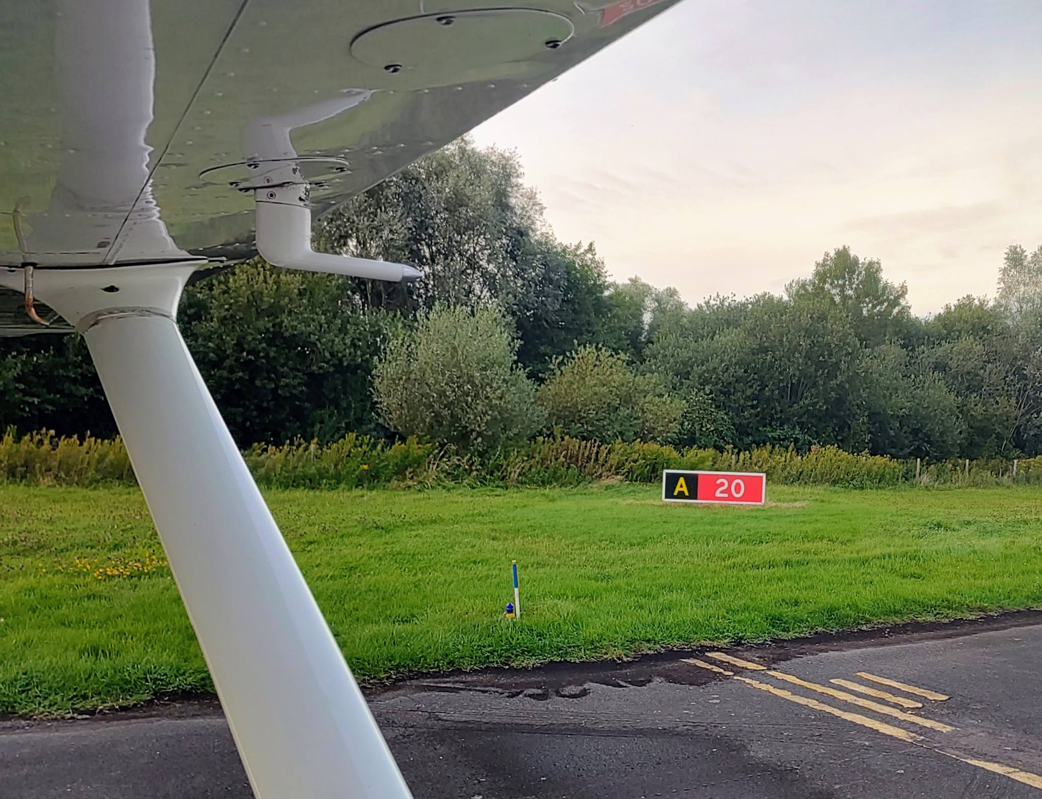 view from the cockpit of a Cessna C172, on the taxiway, looking to the left; yellow markings of a holding point can be seen, with the holding point sign "A 20" in the background