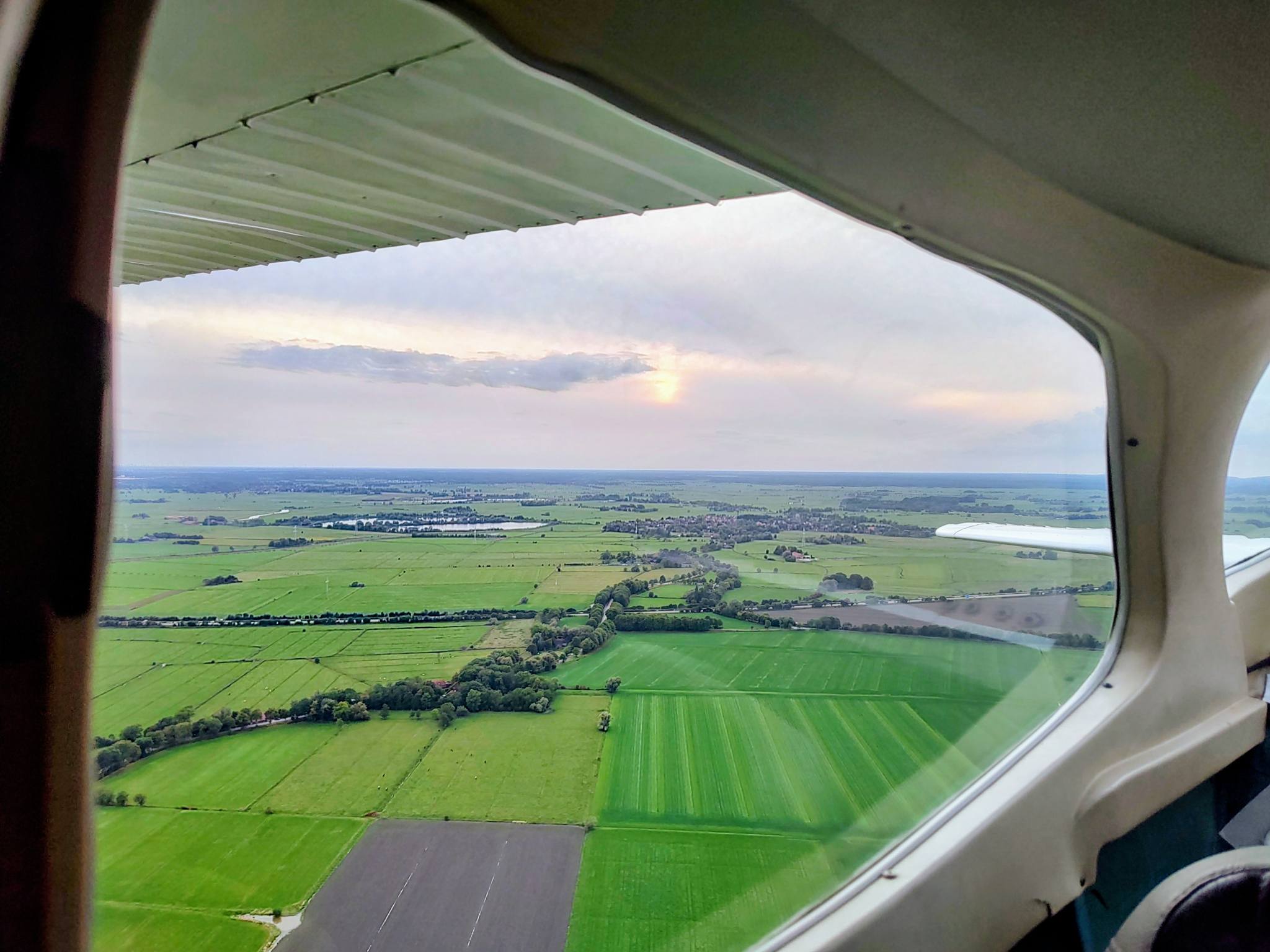 view from the cockpit of a Cessna C172 flying at 800 ft, looking back to the right; the setting sun is visible behind clouds/haze, below are green meadows and lines of trees.