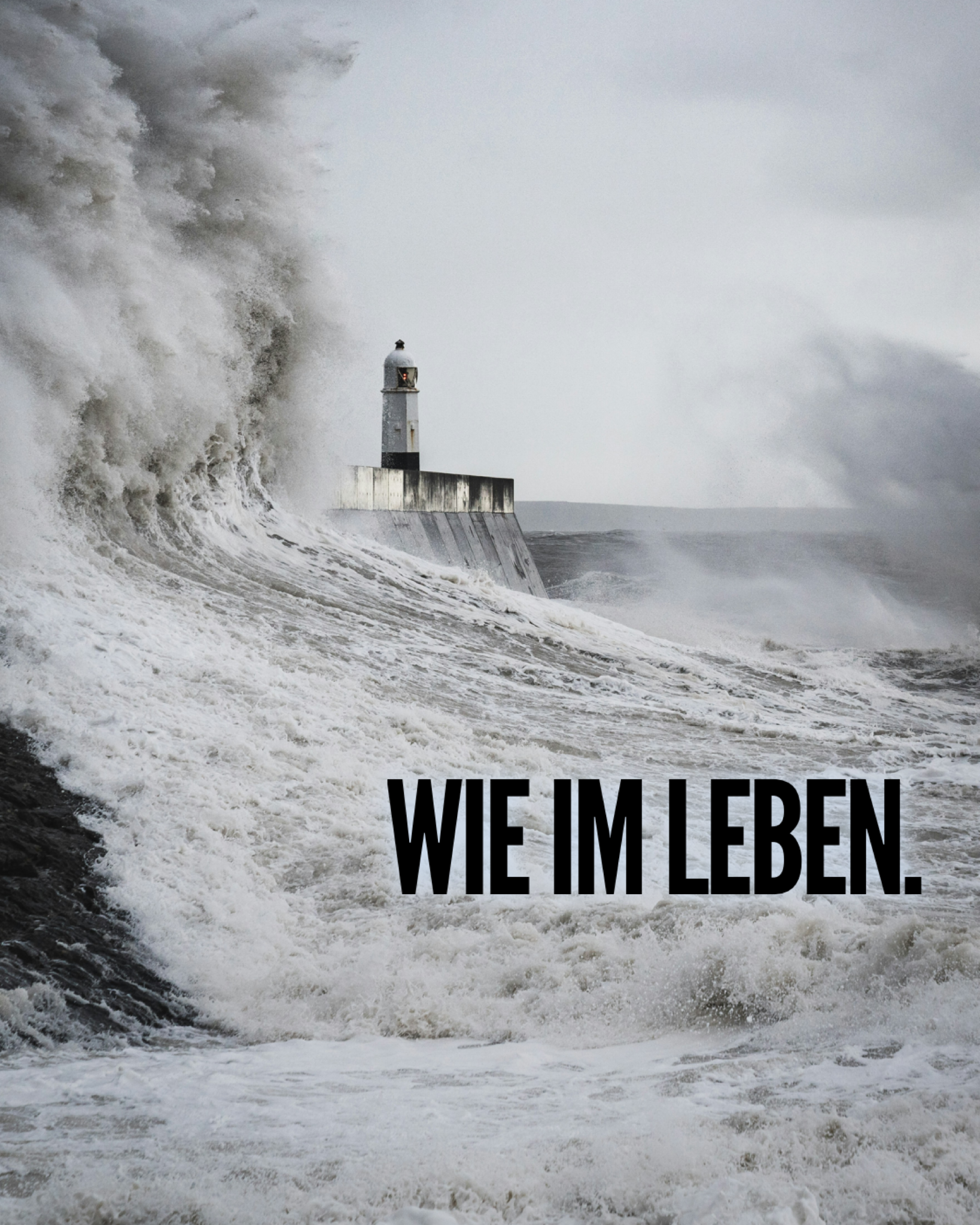 Hohe, grauweiße Sturmwellen schlagen gegen eine Mole, auf der ein kleiner Leuchtturm steht. Das Meer ist aufgewühlt, Gischt spritzt in die Höhe, und der Himmel darüber ist dicht bewölkt und grau.