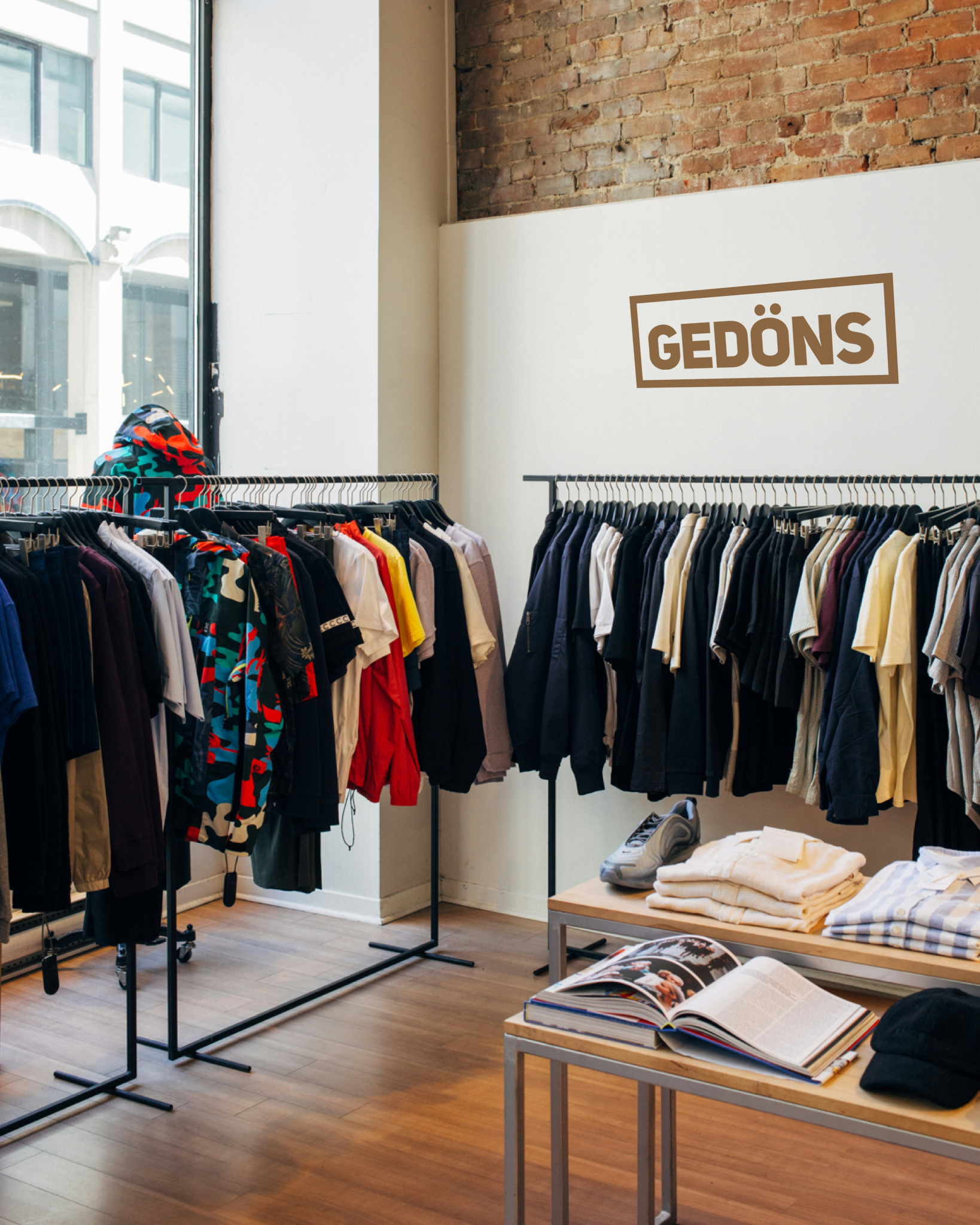 Innenaufnahme eines kleinen Modegeschäfts mit hellen Wänden und freigelegter Backsteinwand. Links und rechts stehen zwei lange Kleiderstangen voller bunt gemischter Kleidung, überwiegend T-Shirts, Hemden und Jacken. Im Vordergrund stehen zwei niedrige Tische mit gefalteten Pullovern, einem Paar Sportschuhe, einem geöffneten Bildband und einer dunklen Kappe. Durch das große Schaufenster links fällt Tageslicht in den Raum.