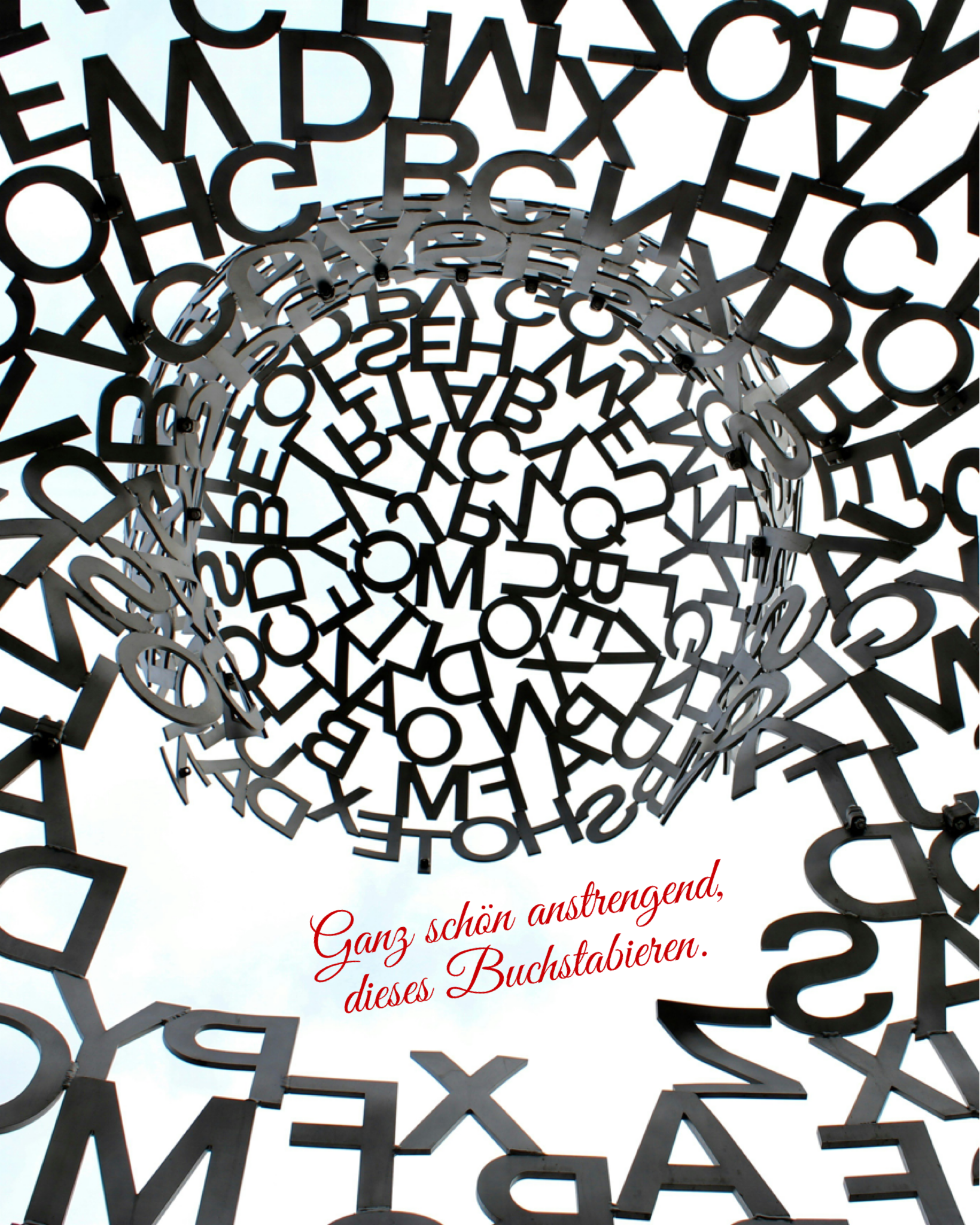 Blick von unten in eine skulpturartige Konstruktion aus schwarzen, überlappenden Großbuchstaben, die ringförmig einen offenen Kreis nach oben bilden. Durch die Zwischenräume ist der helle Himmel zu sehen.
