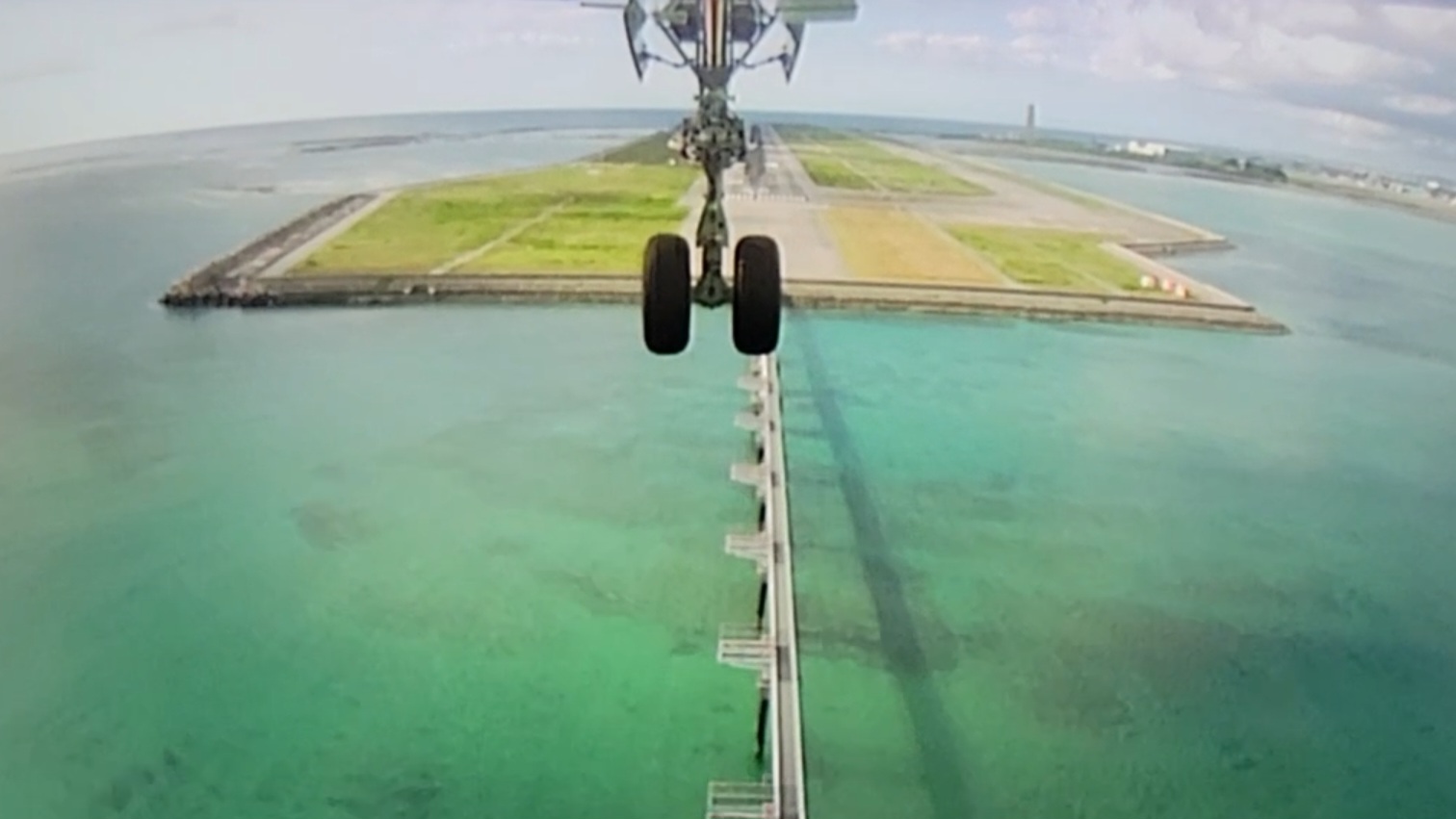 A view from an aircraft's landing camera as it approaches a runway built on a man-made island. The aircraft's landing gear is visible in the center of the image, descending toward the clear turquoise water below. The runway stretches out ahead, surrounded by the ocean, with a coastal landscape and buildings faintly visible in the distance under a partly cloudy sky.