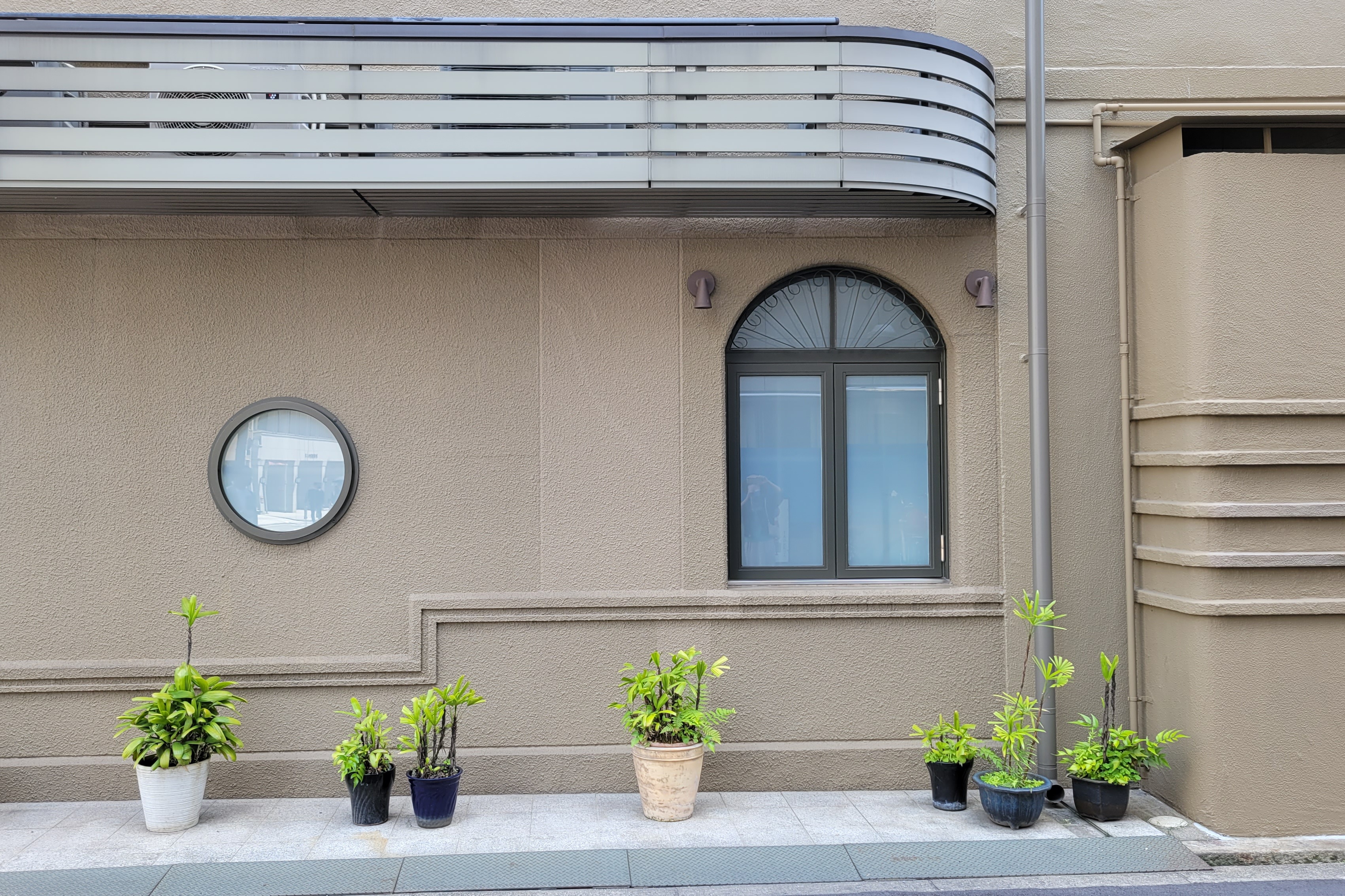 A beige exterior wall with a circular window on the left and an arched window with frosted glass on the right. Above the arched window is a curved slatted covering. In front of the wall are seven potted green plants of various sizes and containers, neatly arranged along the sidewalk.