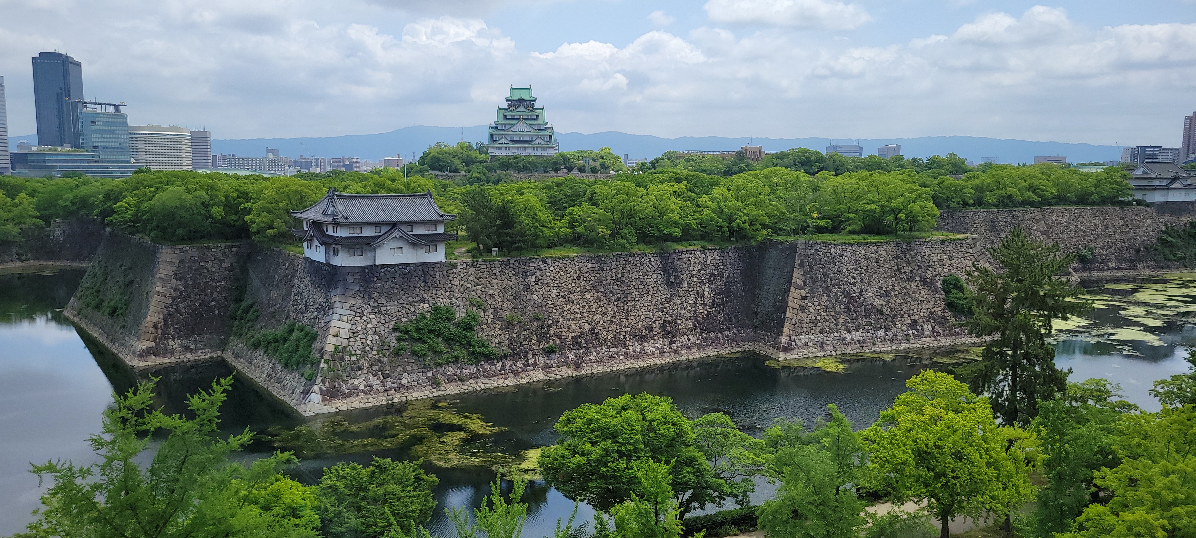 A panoramic view of Osaka Castle in Japan, surrounded by lush green trees and a broad stone moat. The central tower of the castle, with its iconic green roof and white walls, rises above the treetops. The massive stone walls of the castle grounds are clearly visible, with a traditional watchtower at the corner. A calm body of water reflects parts of the scenery, and modern buildings from Osaka’s cityscape are visible in the background, blending historical and contemporary elements. The sky is partly cloudy with soft lighting.