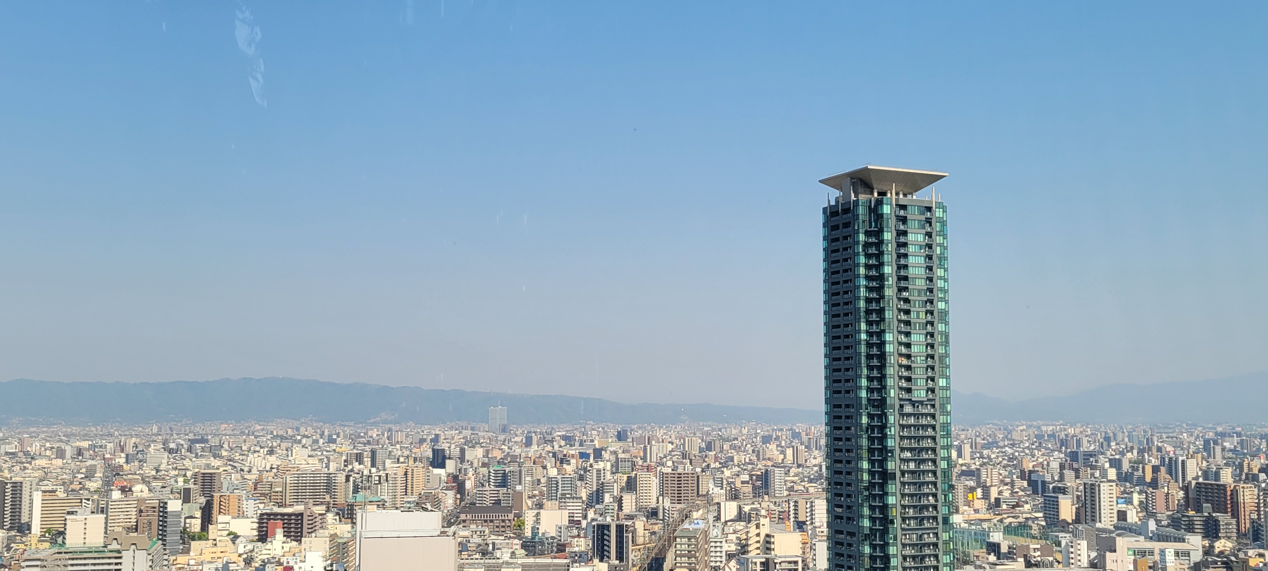 Panoramic view of a sprawling urban cityscape under a clear blue sky. A tall, modern, green-glass skyscraper with a distinctive square roof structure stands prominently in the foreground on the right side of the image. Countless shorter buildings spread across the city below, with distant mountains lining the horizon.
