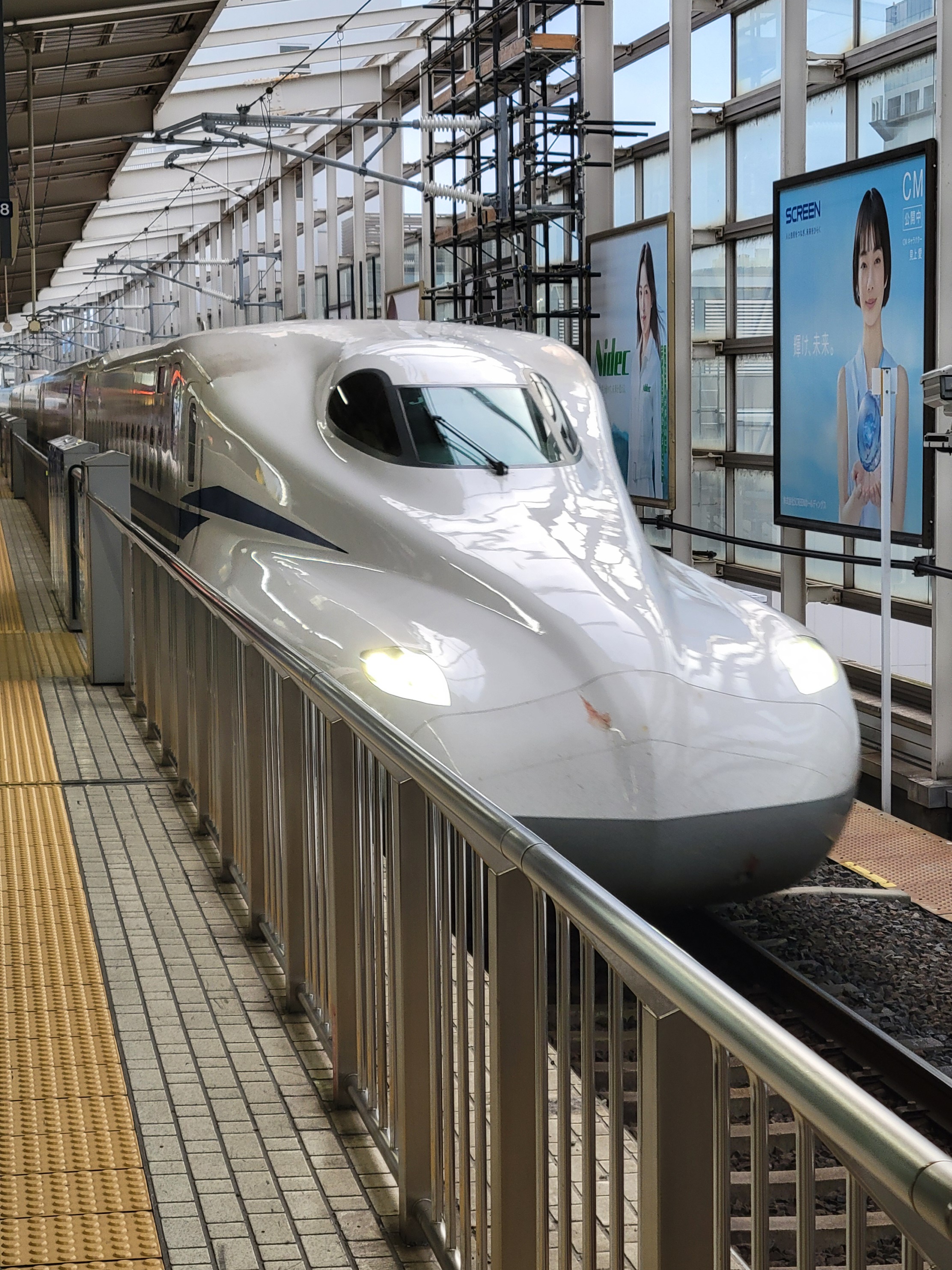 A white N700 series Shinkansen bullet train is arriving at an elevated platform of Kyoto Station in Japan. The train's aerodynamic nose is prominently featured. Overhead, there are power lines and structural beams, and on the right side of the image are advertising posters with Japanese models promoting various products.