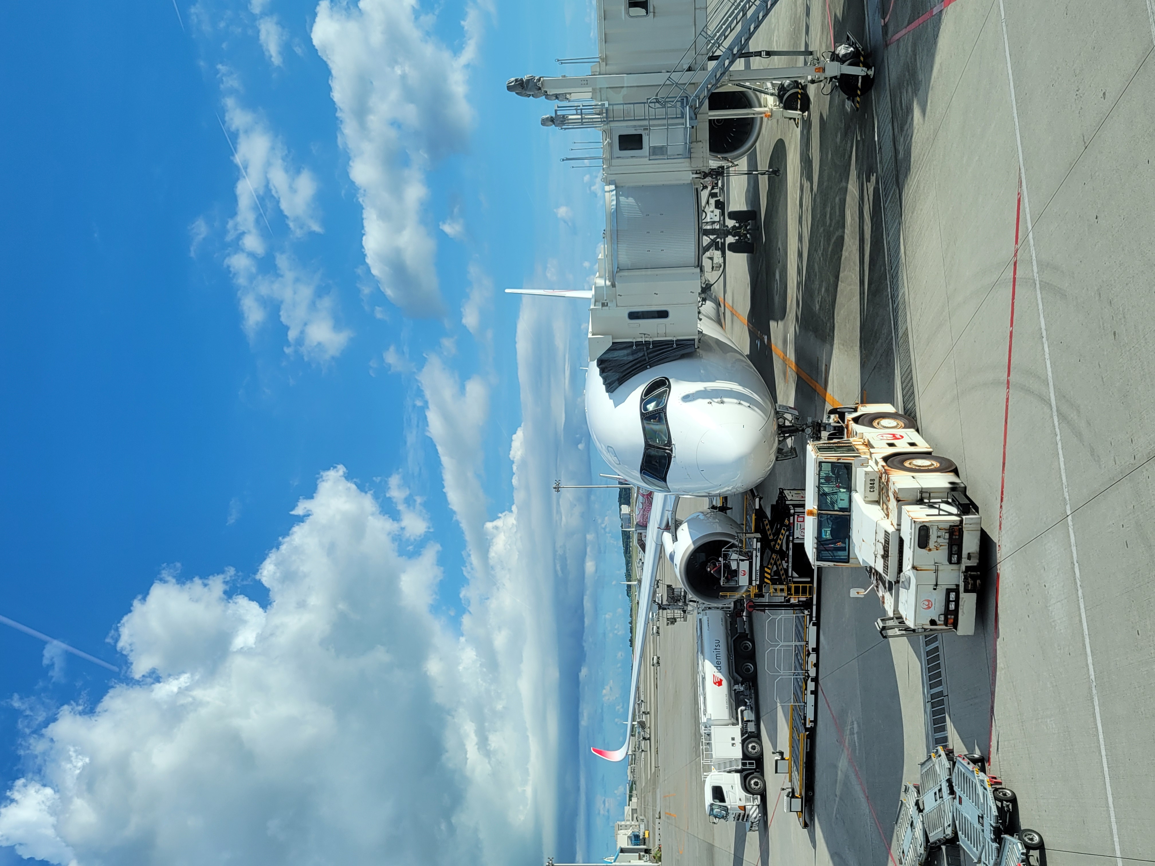 A commercial jet is parked at an airport gate under a bright blue sky with scattered clouds. The aircraft is connected to a passenger boarding bridge and surrounded by ground service vehicles, including a pushback tug in the foreground and fuel and baggage trucks nearby. The plane's sleek nose and cockpit windows are prominently visible, with a clear view of the airport tarmac and runway area in the background.