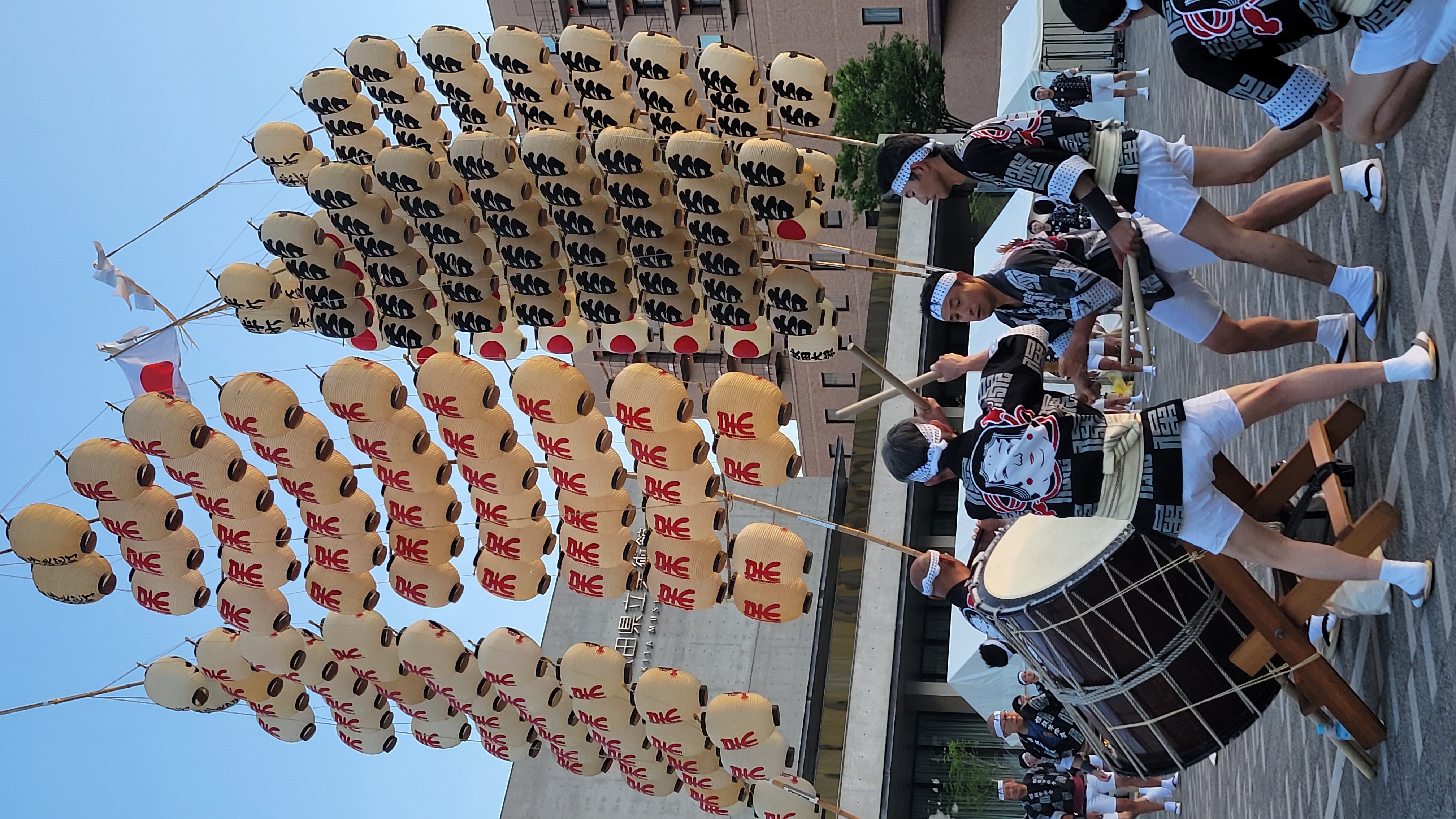 A scene from the Kanto Matsuri festival in Akita, Japan. Multiple performers in traditional black and white festival attire balance enormous bamboo poles adorned with rows of paper lanterns. The lanterns, decorated with bold red and black kanji characters, sway against the blue evening sky. In the foreground, several men play a large taiko drum. The coordinated performance takes place on a public plaza, with spectators and other performers in the background.