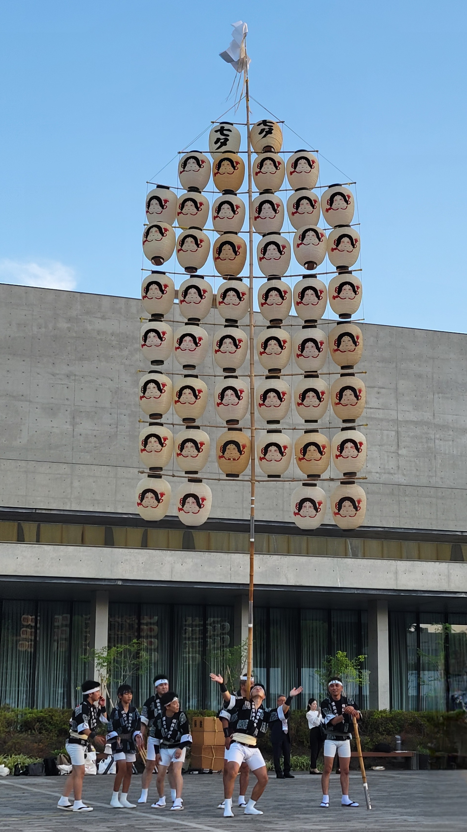 A group of performers participate in the traditional Kanto Matsuri festival in Akita, Japan. One performer balances a tall bamboo pole on his shoulder, supporting a large horizontal array of illuminated paper lanterns. The lanterns are decorated with stylized faces of women and arranged in tiers, forming a fan shape. Other members of the group, dressed in matching black happi coats and white shorts, stand nearby in support. The performance takes place outdoors against a modern concrete building, with a clear blue sky above.