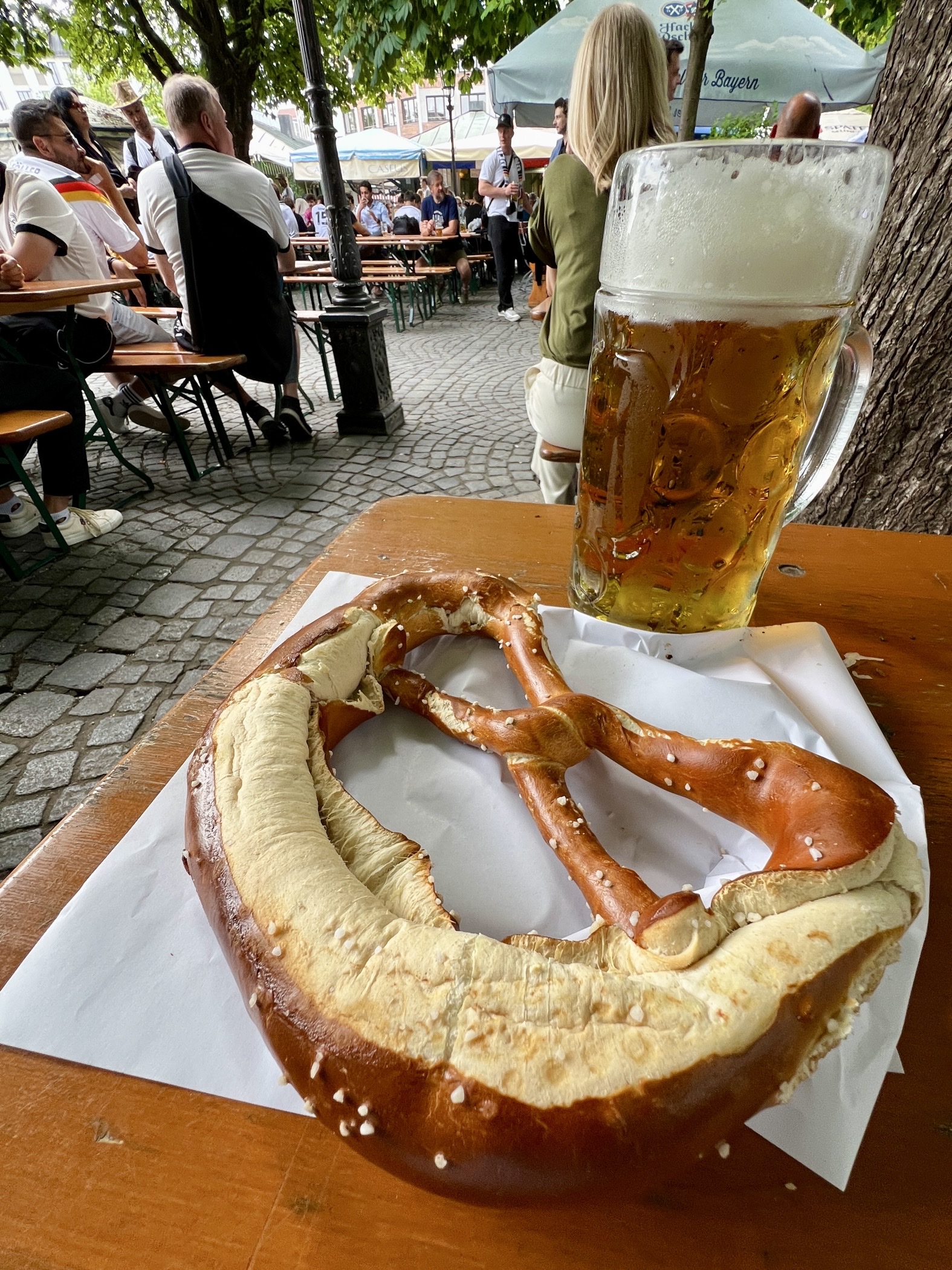 hochkant weitwinkliges Foto eines Biertischs mit einer Maß Bier und einer großen Brezn, andere Menschen im Hintergrund