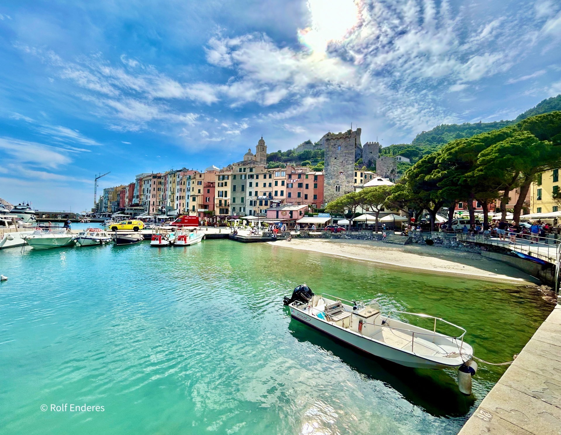 A scenic harbor lined with colorful buildings, boats docked in calm turquoise water, and a backdrop of rolling hills under a partly cloudy sky. A beach and people can be seen enjoying the lively atmosphere.