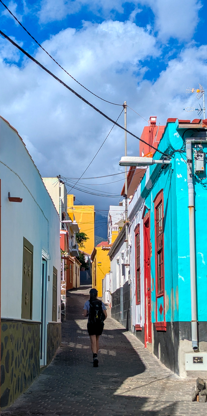 Das Bild zeigt eine Person mit einem Rucksack, die auf einer engen Gasse zwischen farbenfrohen Gebäuden hindurchgeht. Die Häuser sind in verschiedenen Farben wie Weiß, Gelb und Türkis gestrichen, was der Szene eine lebendige und malerische Atmosphäre verleiht. Der Himmel ist blau mit einigen Wolken, und die Sonne scheint und wirft Schatten auf die Gasse.