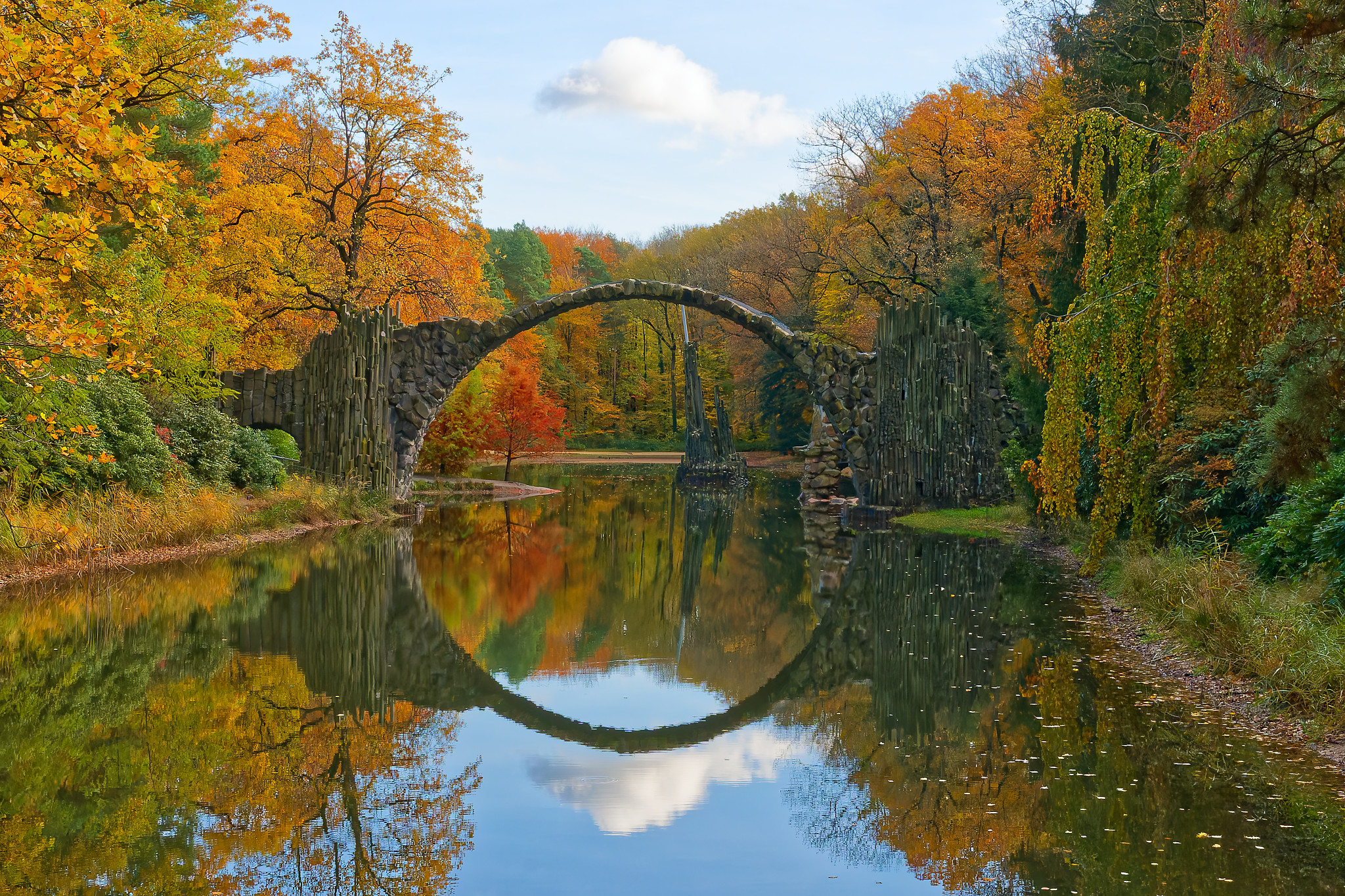 Steinerne Bogenbrücke über einen Teich, umgeben von herbstlich bunt gefärbtem Laub an Bäumen.