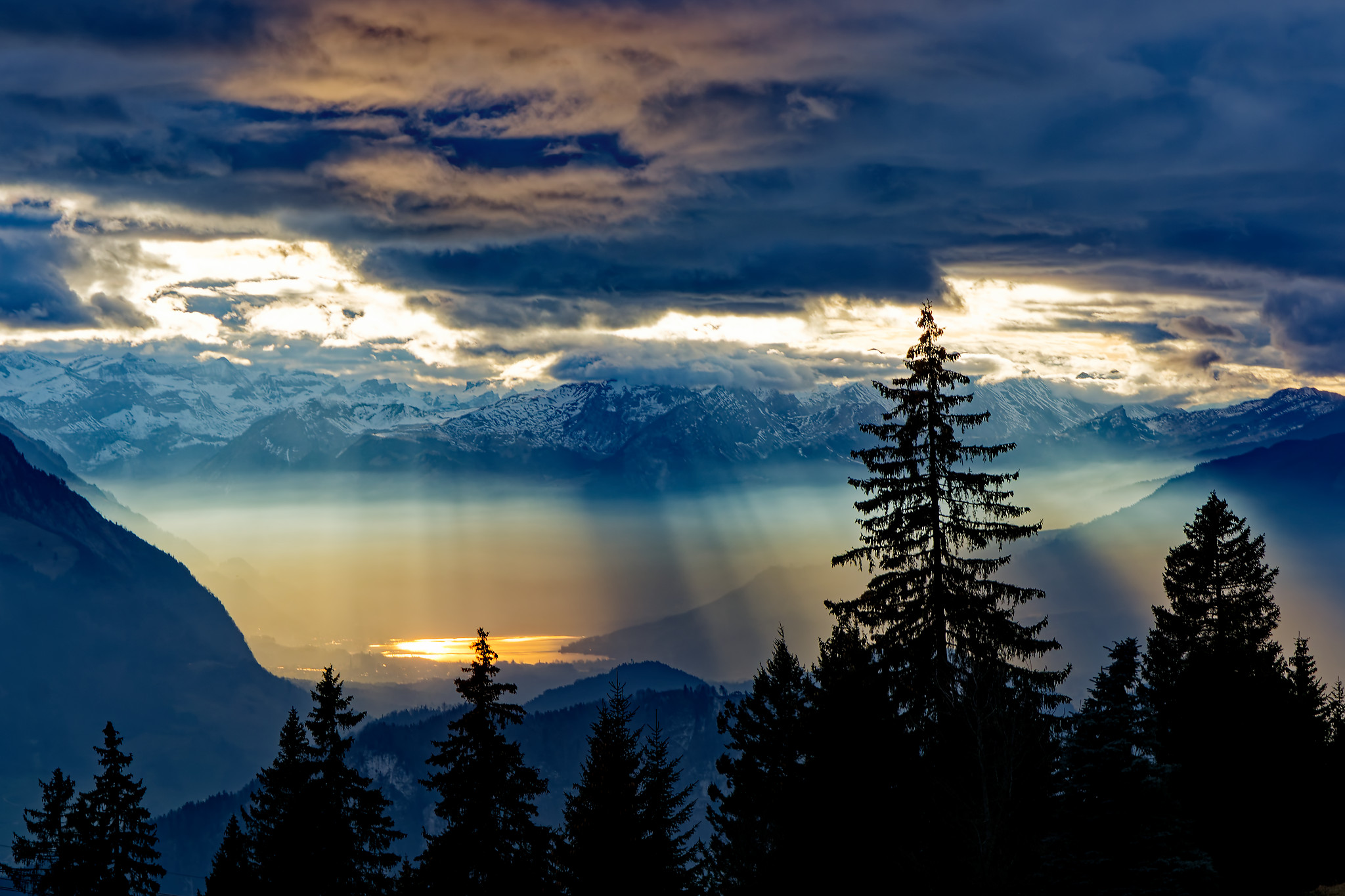 Dunkle Silhouetten von Tannenbäumen im Vordergrund vor einem Panorama aus Bergen und einem See, der von Sonnenstrahlen durch Wolkenlücken beleuchtet wird. Der Himmel zeigt dramatische Wolkenformationen mit hellen und dunklen Bereichen, die das Lichtspiel verstärken.