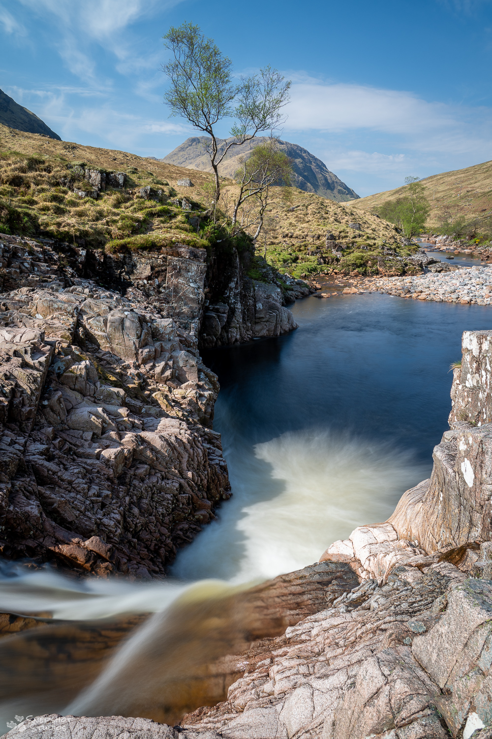 Photo looking down-river over a small waterfall in a valley in Scotland. a single tree grows on the cliff above the river.