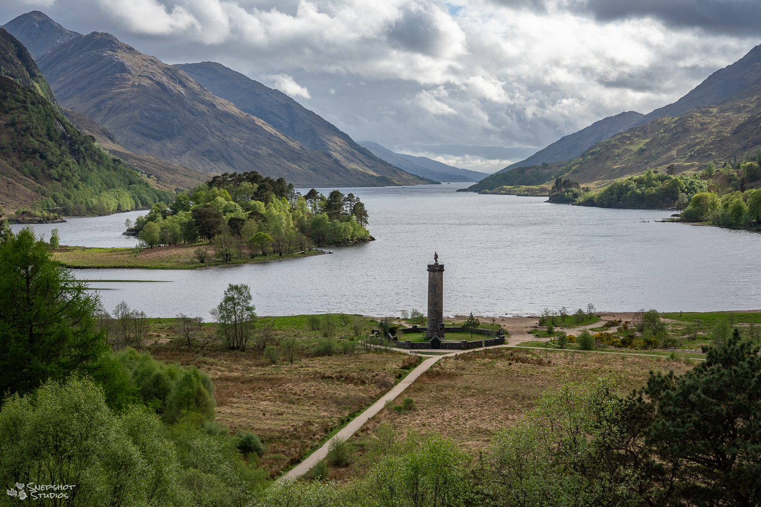 view along a narrow Loch in Scotland. in the foreground, at the end of the Loch, stands a column as a monument