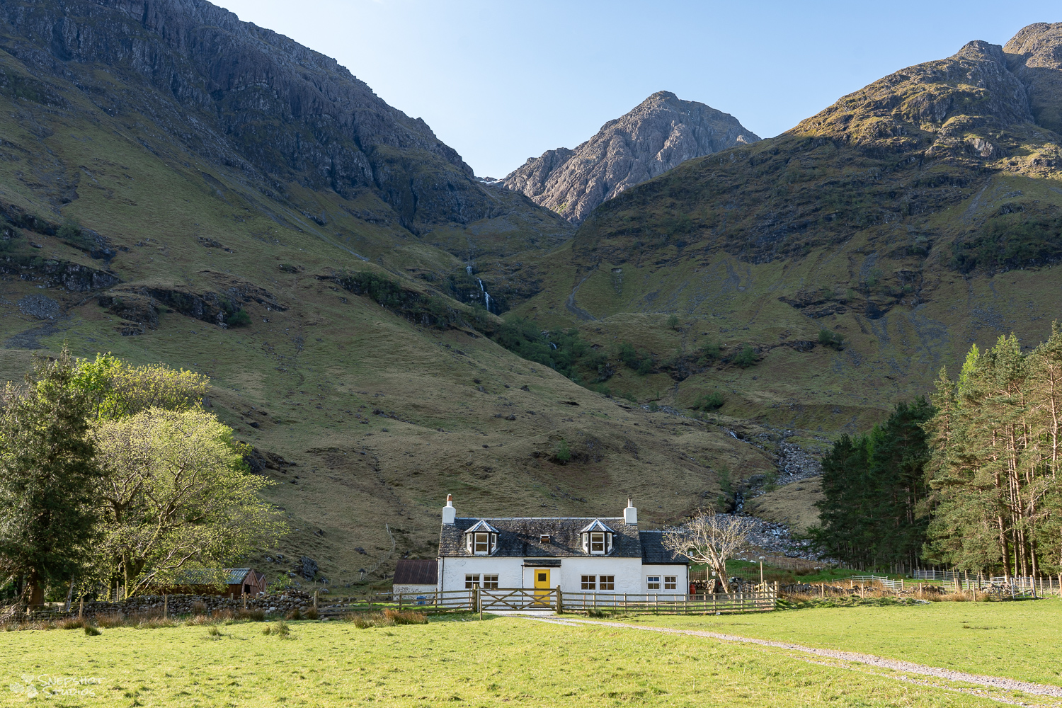 a small white house with a yellow door. Flanked on both sides by trees and with a backdrop of impressive mountains