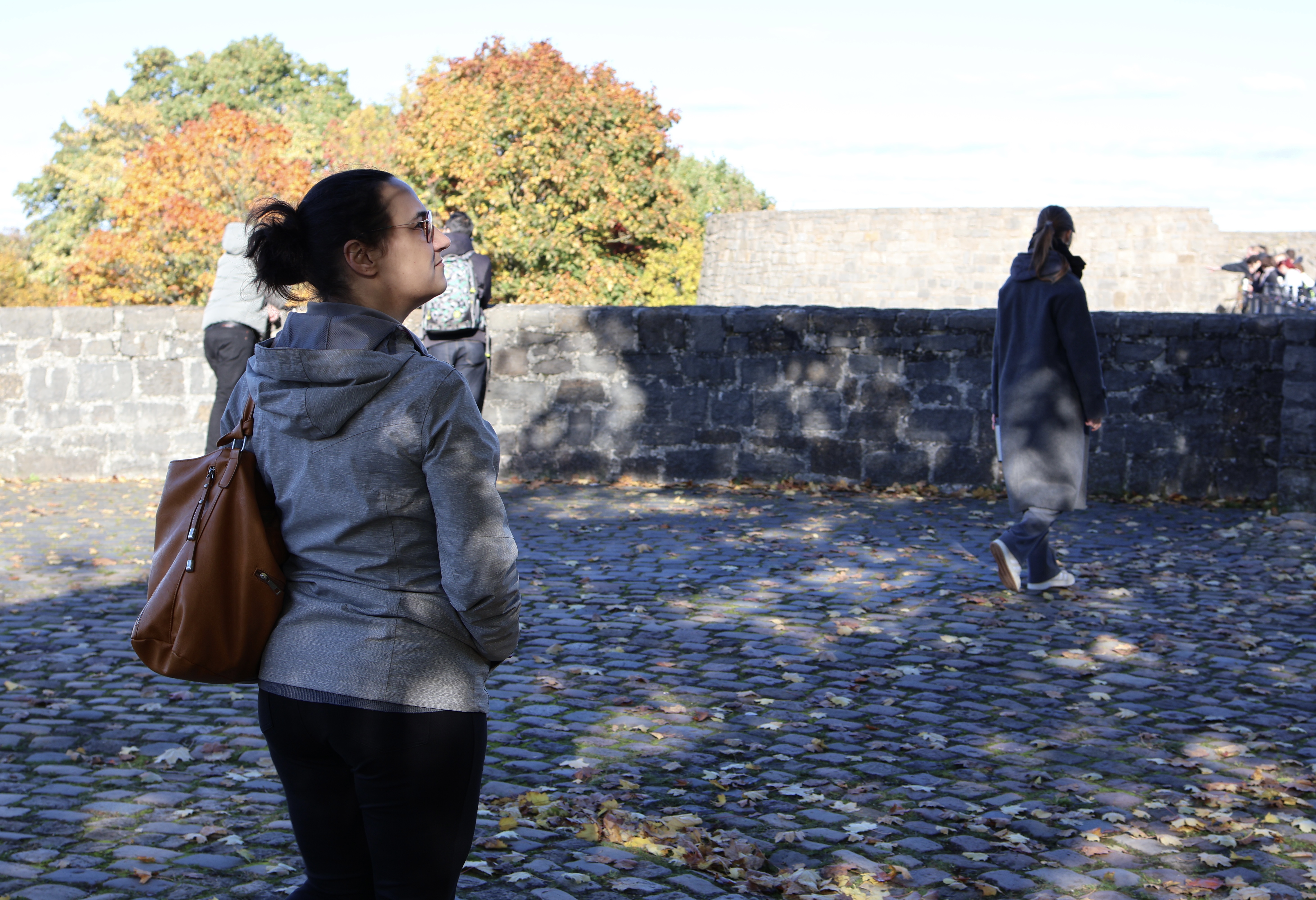 Ein Platz auf der Sparrenburg mit Kopfsteinpflaster. Drumherum eine schulterhohe Mauer aus Bruchsteinen, dahinter Laubbäume und ein Dachfirst. Nahe der Mauer geht eine Frau, man sieht sie von hinten. Im linken Vordergrund des Bildes steht Frau K., ebenfalls in Rückenansicht, aber den Kopf so gedreht, dass man ihr Gesicht im Profil sehen kann. Sie hat eine graue Jacke und eine schwarze Hose an. Über der linken Schulter trägt sie eine große, braune Ledertasche.