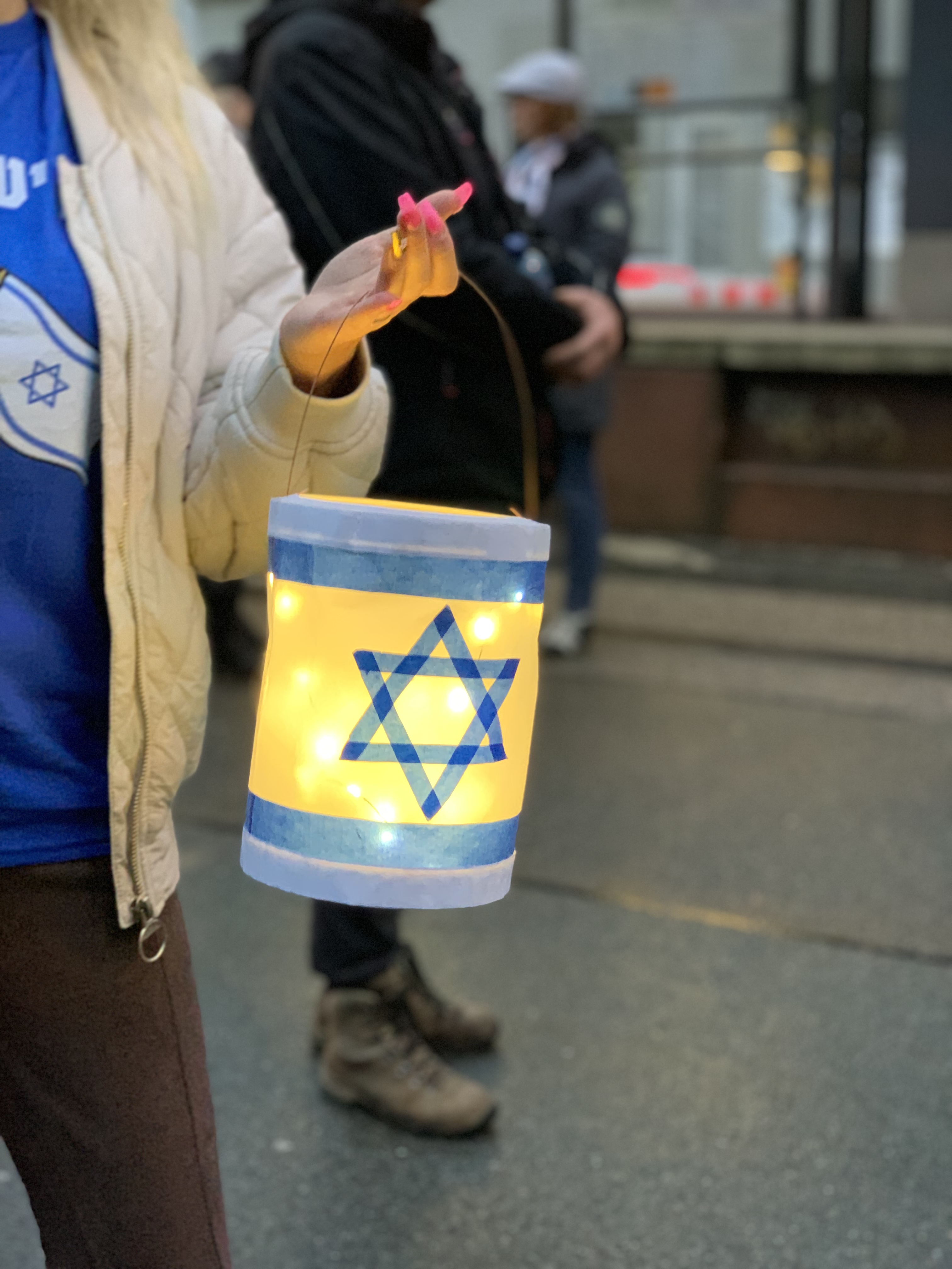 Eine selbst gebastelte Laterne mit der Flagge Israels als Motiv. Gehalten von einer Frauenhand mit langen pinken Fingernägeln, der Ringfinger aber in golden. Sie trägt außerdem ein Israel-T-Sgirt unter der Jacke und eine braune Hose. Bildausschnitt ohne Kopf. Im Hintergrund verschwommen zwei weitere Teilnehmer und die Kante der Straßenbahnhaltestelle gegenüber der Synagoge.