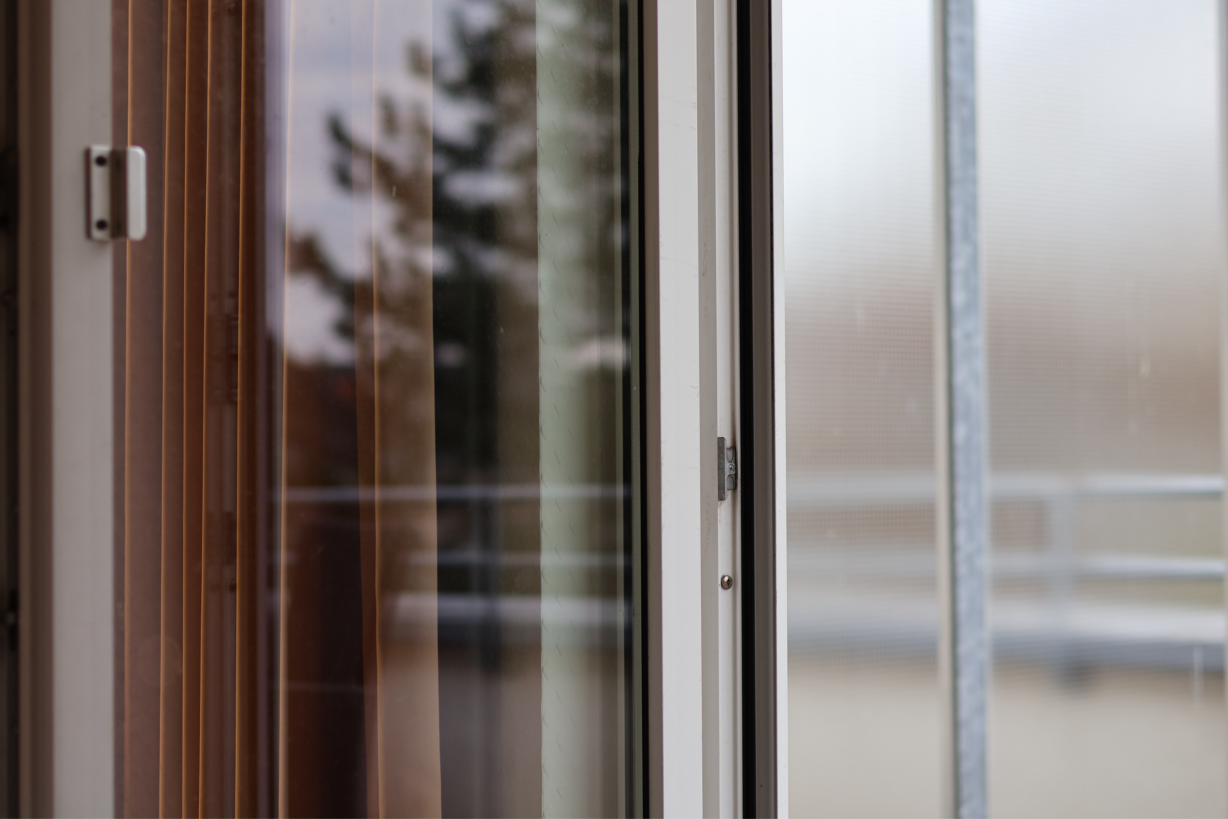 Links der Rahmen der Balkontür mit Griff, dahinter etwas brauner Vorhang, dann Fensterglas, in dem sich ein Baum spiegelt, dann der verschwommen3 Blick nach draußen bei diesigem Wetter.