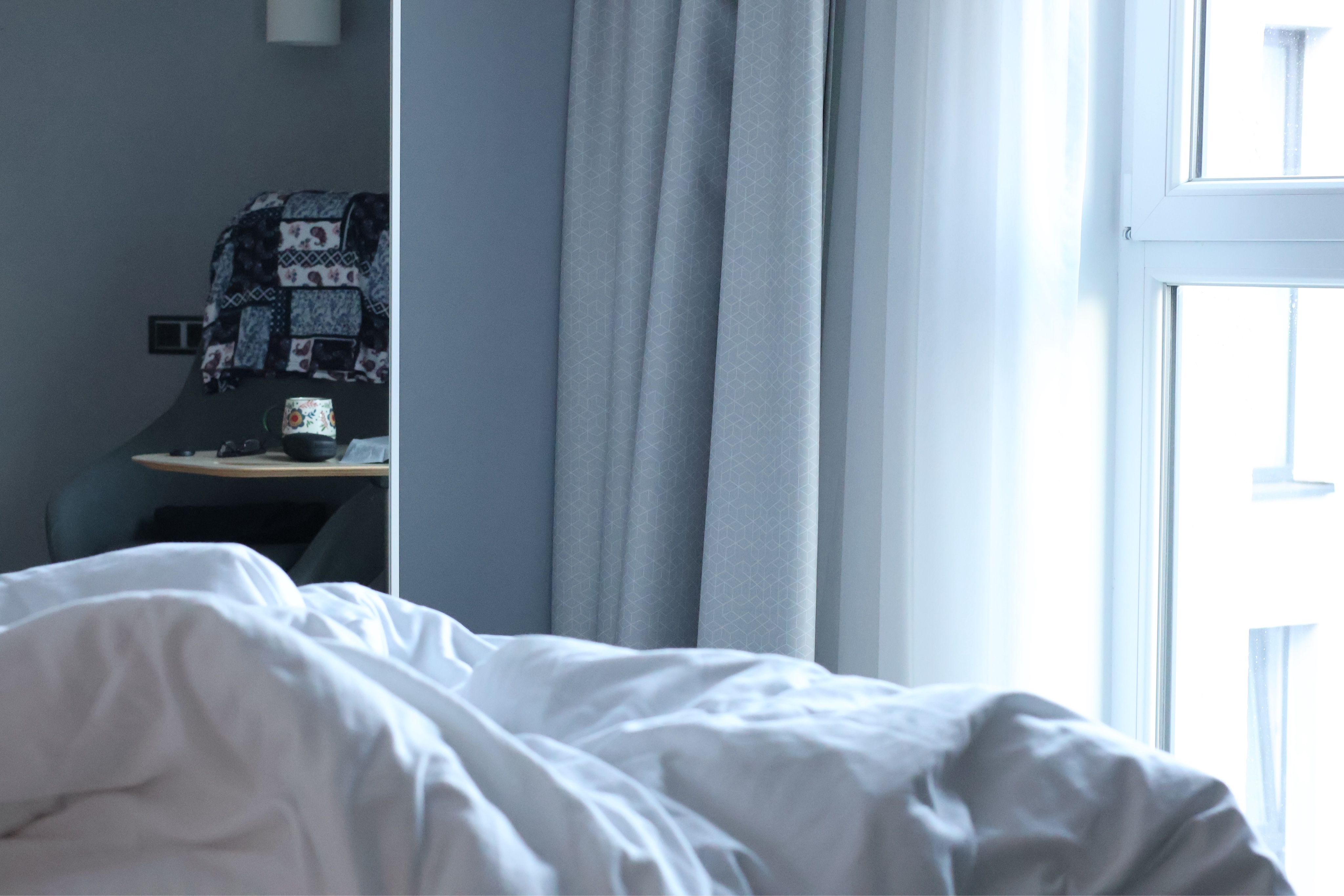 Im Vordergrund die zerwühlte Bettdecke, weiß bezogen. Dahinter sieht man links in einem wandhohen Spiegel einen Sessel mit einem gemusterten Shirt überder Lehne. Auf dem Tisch davor steht ein Teepott, daneben liegen ein paar kleinere Sachen. Rechts daneben in der Bikdmitte die hellgraue Wand, dann ein hellgrauer Vorhang, dann eine weiße, zur Seite geschobene Gardine. Endlich das Fenster mit weißem Licht.