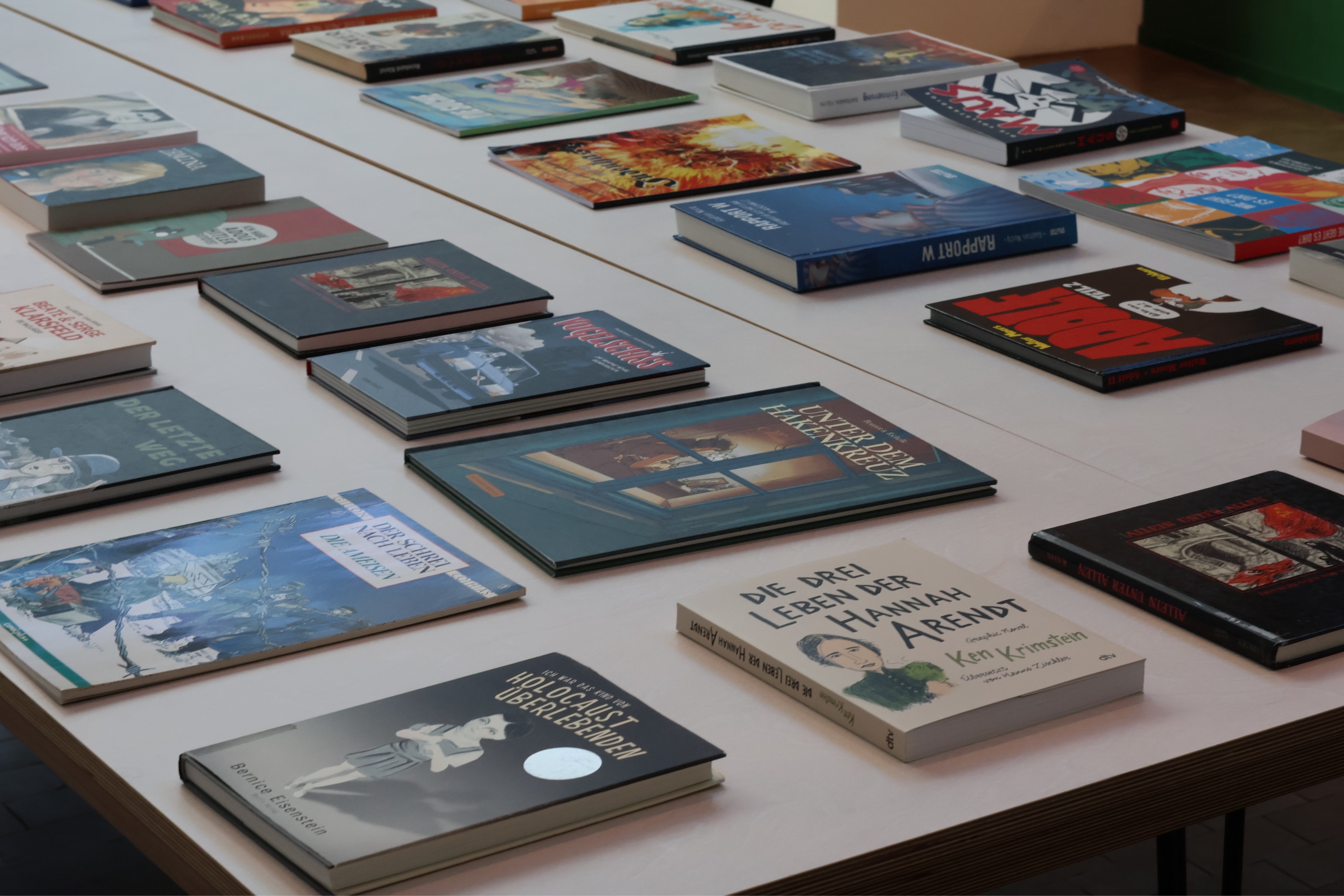 Teil des Tisches (man sieht, dass er aus mehreren zusammengeschoben ist) mit vielen bunten Büchern nebeneinander.