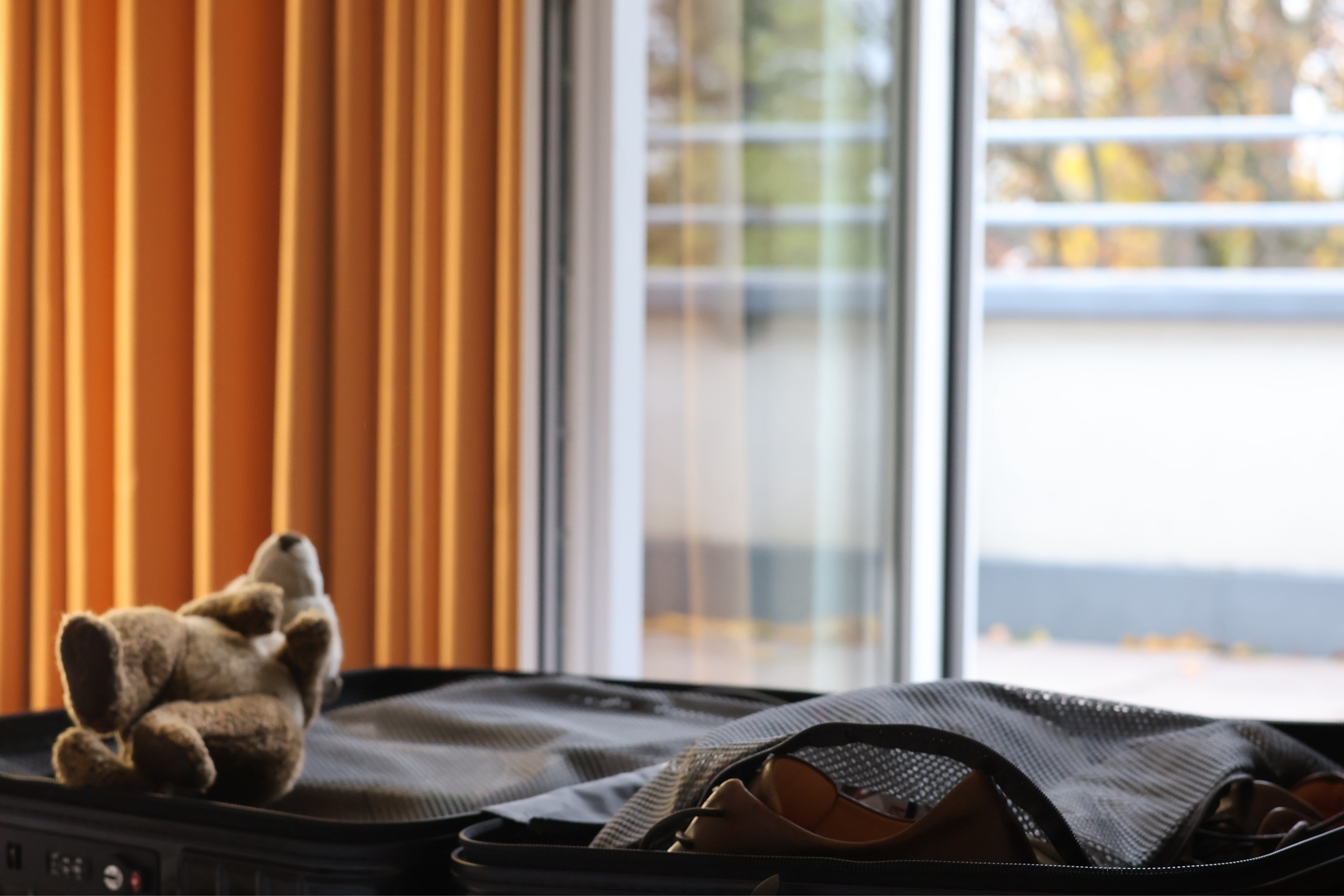 Wieder der Blick Richtung Balkon, dreigeteilt in Vorhang, Fensterglas und offner Balkontür. Davor ein aufgeklappter Rolkoffer, auf dem links ein Stofferdmännchen mit den Füßen Richtung Betrachterin liegt.