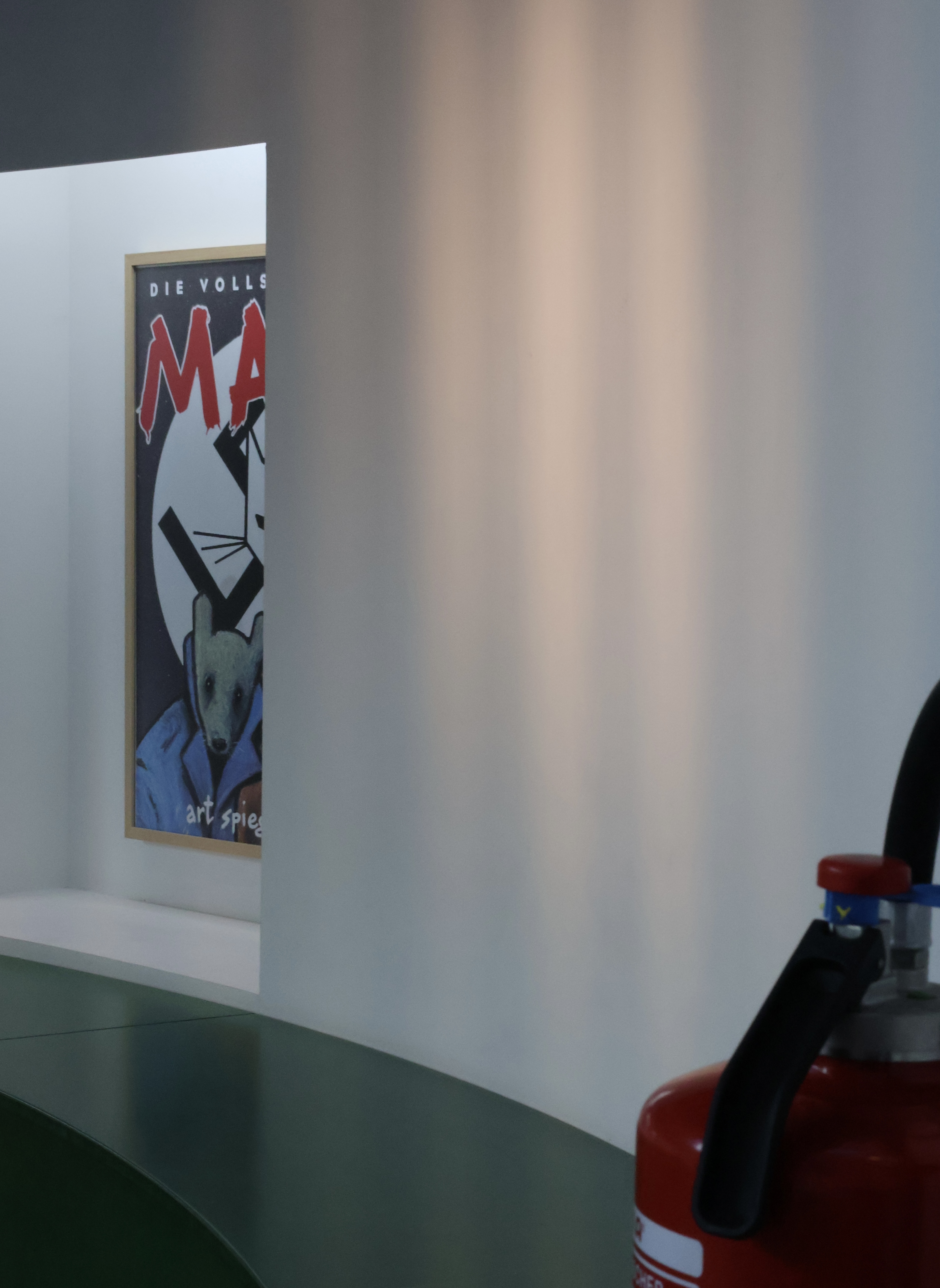 Im Hintergrund eine halbrunde Nische mit einer fensterartigen Vertiefung, in der man ein Stück eines Plakates sieht, das das Titelbild der Graphic Novel "Maus" zeigt. Vorne ein Stück eines Feuerlöschers in dem gleichen Rot wie die Überschrift "Maus" der Graphic Novel.