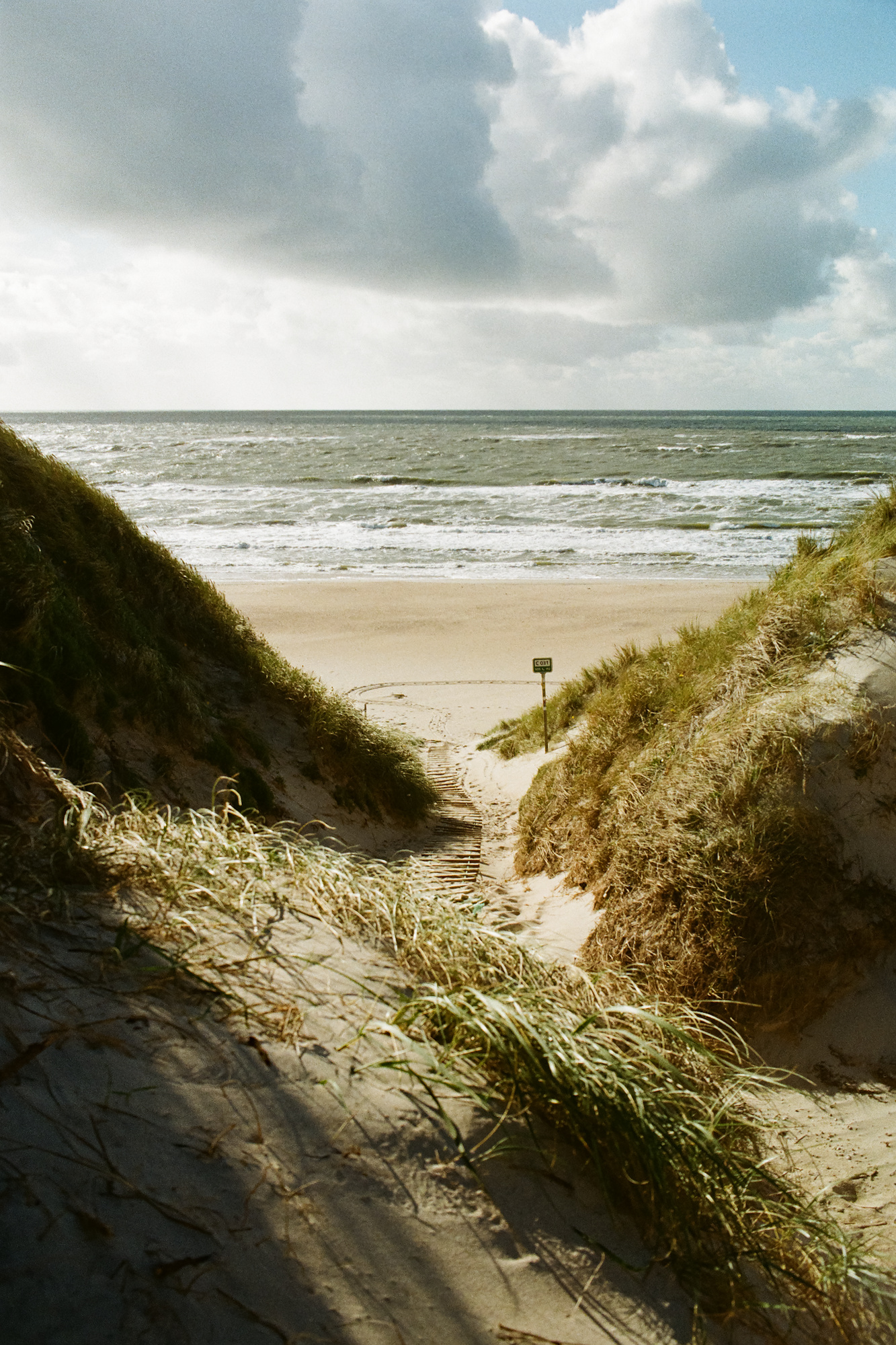 A path leading over a dune to the beach. The sun is shining and there are clouds in the sky.