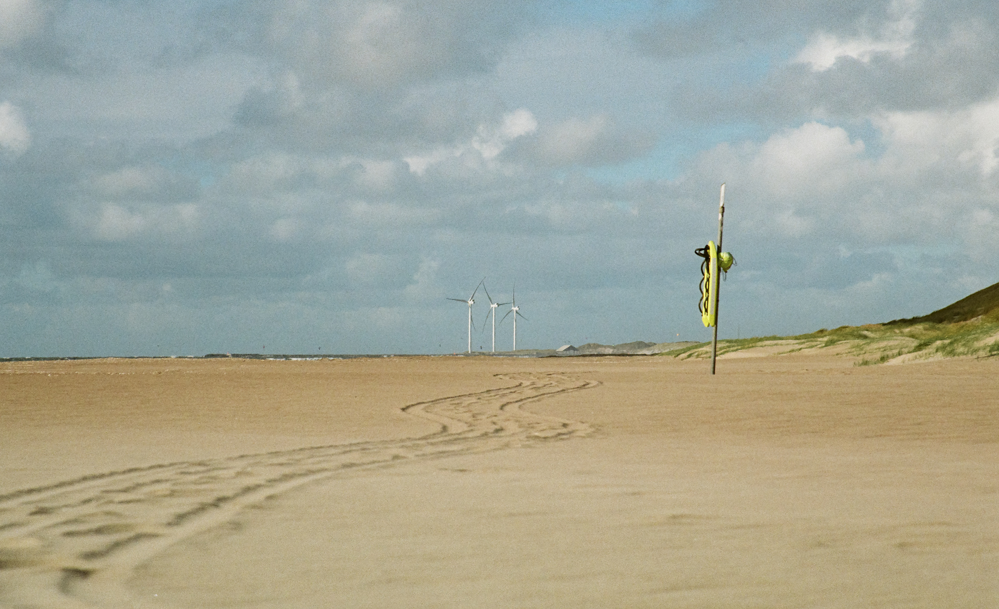 Footprints can be seen in the sand on the beach. In the background there is a lifebuoy and wind turbines.