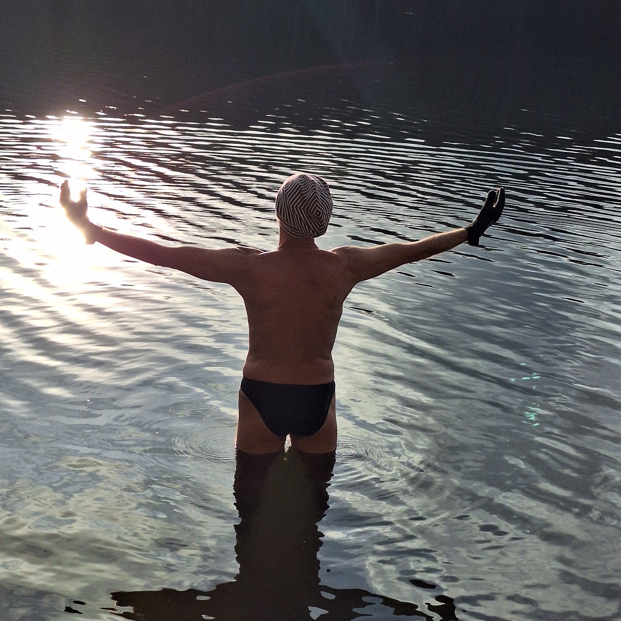 Ein Mann mit schwarzer Badehose und geringelter Mütze steht im Wasser mit ausgebreiteten Armen der Sonne entgegen. Das Sonnenlicht wird auf der leicht gewellten Wasseroberfläche reflektiert.