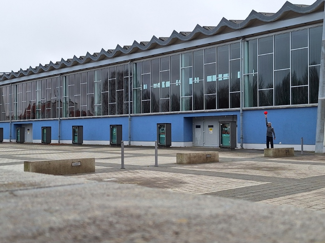 Die langgezogene Fensterfront einer Mehrzweckhalle mit denkmalgeschützter Dachkonstruktion in Fluchtpunktperspektive von rechts nach links aufgenommen. Davor hell- und dunkelgrau gekachelter Steinboden. An der Bildunterkante unscharfe graue Steinfläche. Der Himmel ist wolkenverhangen hellgrau. Als Farbklecks spielt am rechten Bildrand ein Mann mit einem roten Ball vor der hellblauen unteren Gebäudewand.