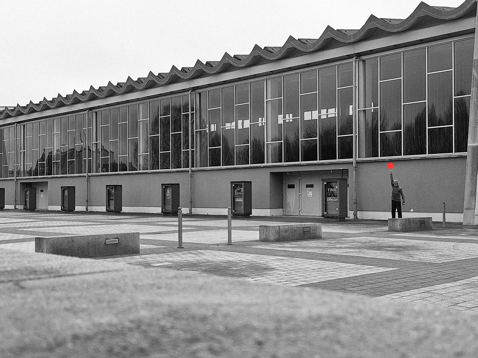 Die langgezogene Fensterfront einer Mehrzweckhalle mit denkmalgeschützter Dachkonstruktion in Fluchtpunktperspektive von rechts nach links in schwarz/weiß aufgenommen. Davor ein gekachelter Steinboden. An der Bildunterkante unscharfe graue Steinfläche. Der Himmel ist wolkenverhangen. Als Farbklecks spielt am rechten Bildrand ein Mann mit einem roten Ball.