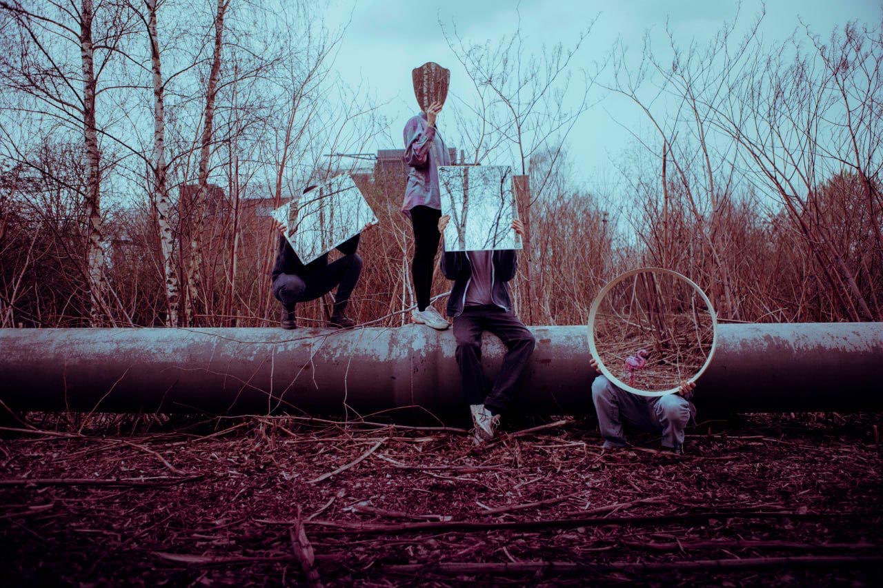 Vier Personen sitzen und stehen auf oder vor einem großen Rohr. Vor ihre Gesichter halten Sie Spiegel.