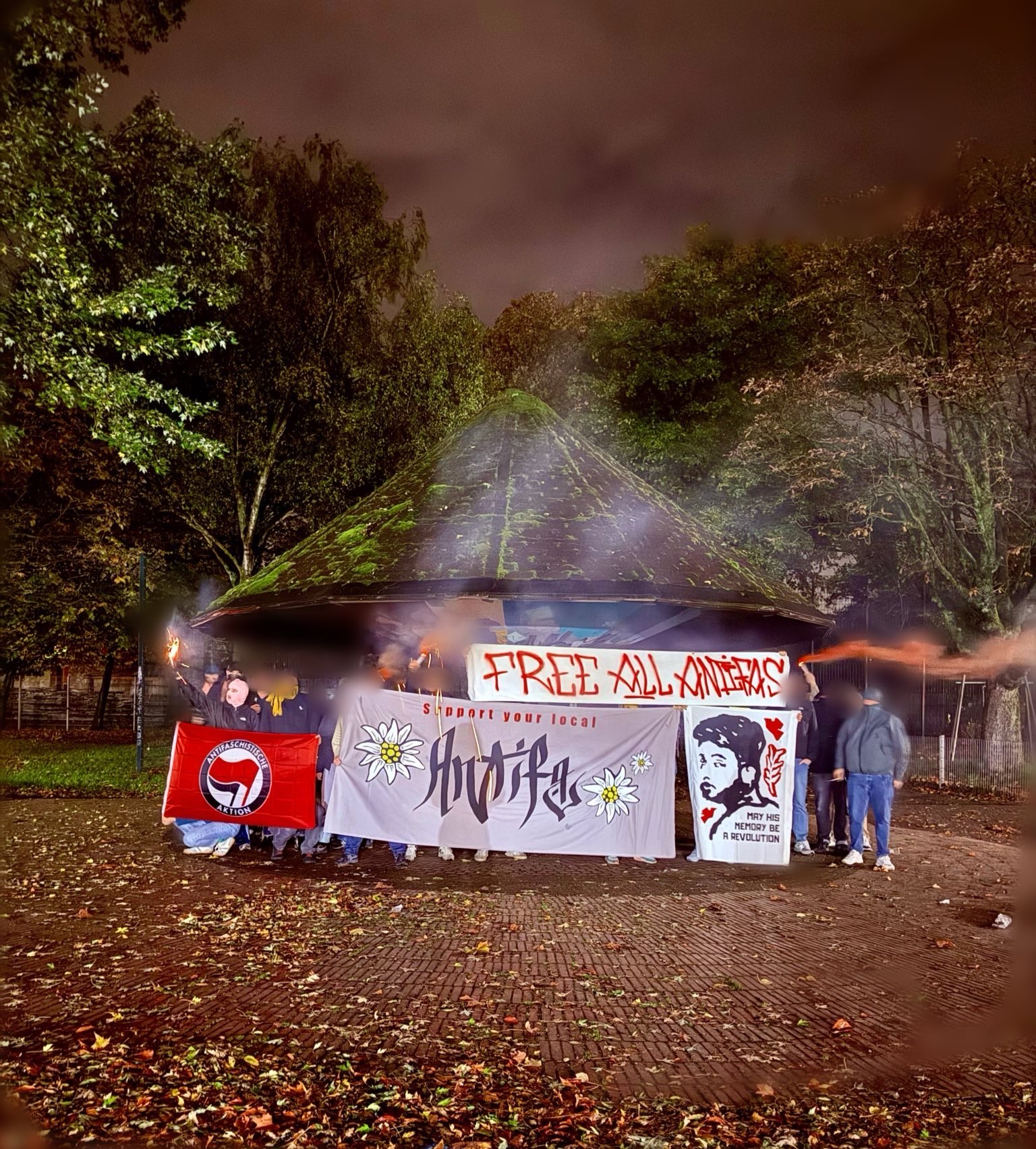 Eine Gruppe Menschen hält Transparente und Pyrotechnike hoch. Zu sehen sind ein Transparent mit der Aufschrift "Support your Local Antifa" neben mehreren Edelweißblüten, ein Transparent mit dem Gesicht von Hersh Goldberg-Polin und der Aufschrift "May your memory be a revolution", eine Tapete auf der "Free All Antifa" gesprüht wurde und eine Antifafahne