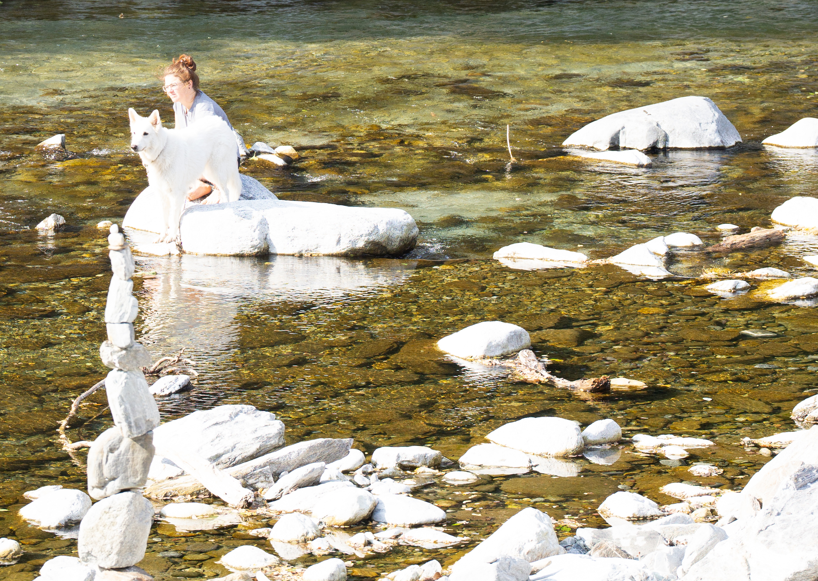 A woman with white clothes sits on a rock beside a white dog in a shallow river, surrounded by smooth white stones and clear water. A balanced white stone tower is in the foreground.